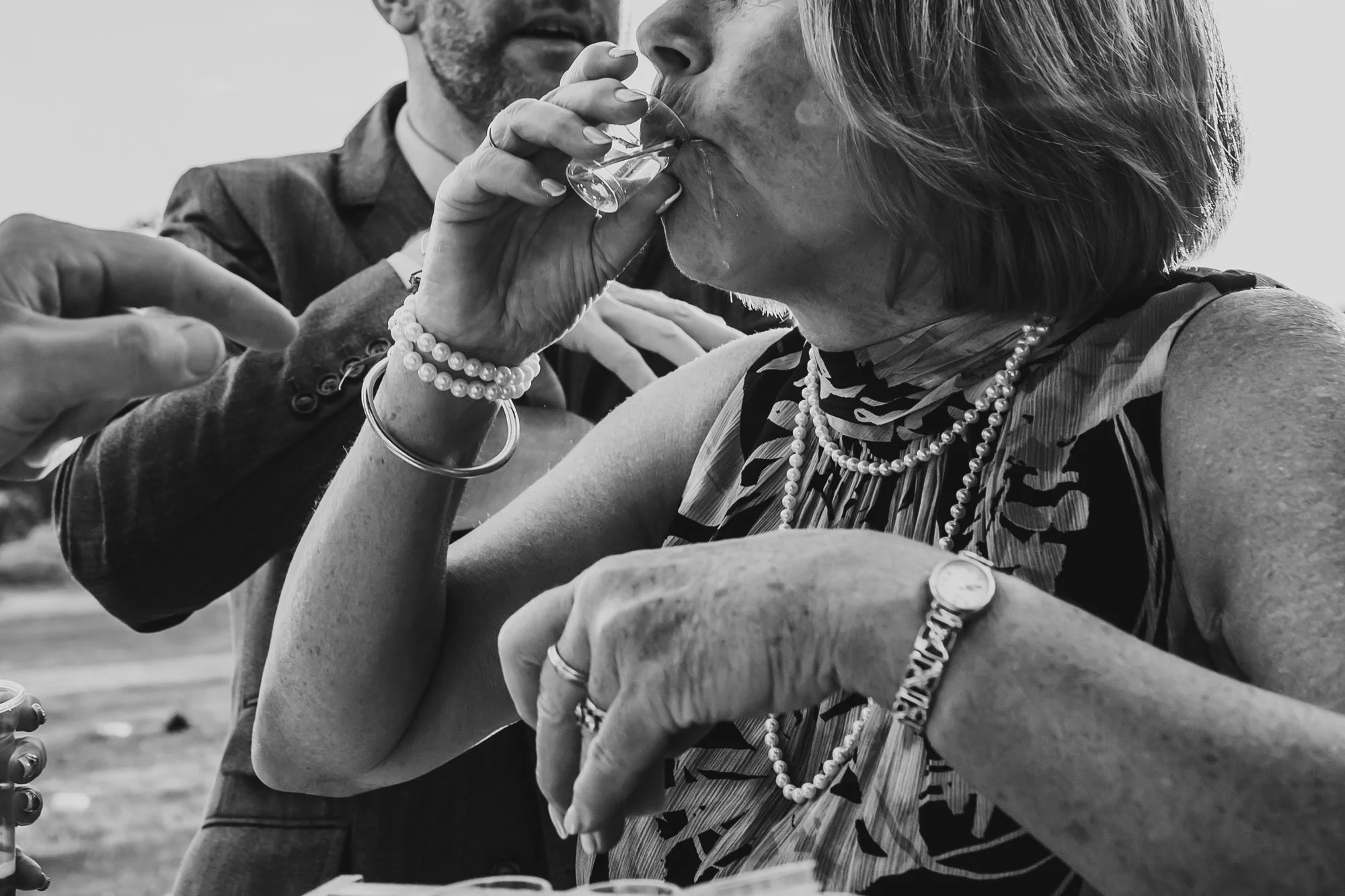 photograph of an older female wedding guest drinking a shot of alcohol. It is dribbling down her chin.