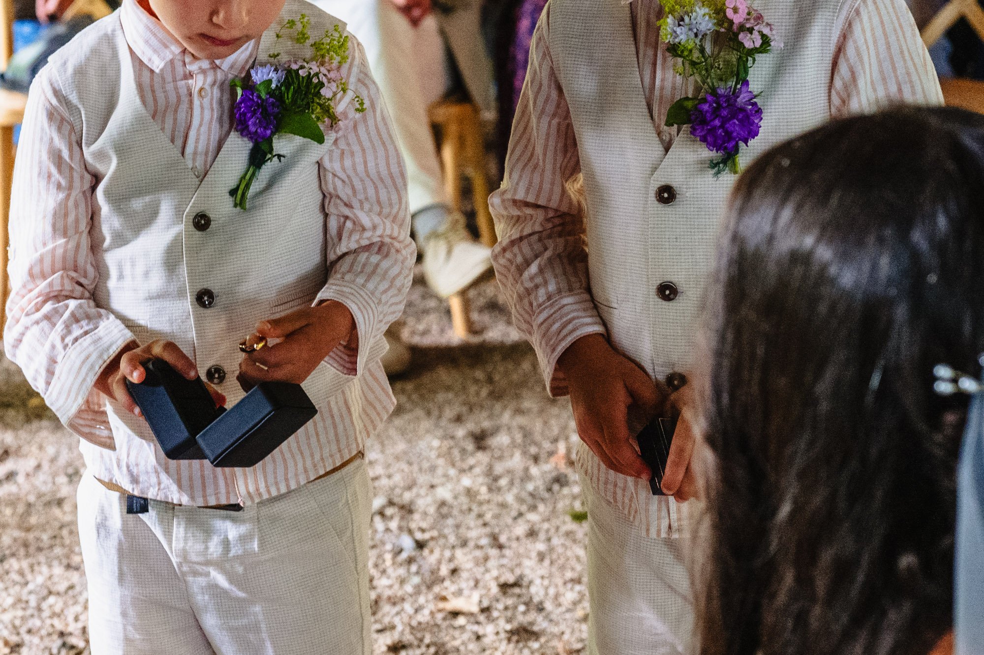 close up image of two young boys wearing summery suits and holding jewellery boxes. One is getting a wedding ring out of a box.
