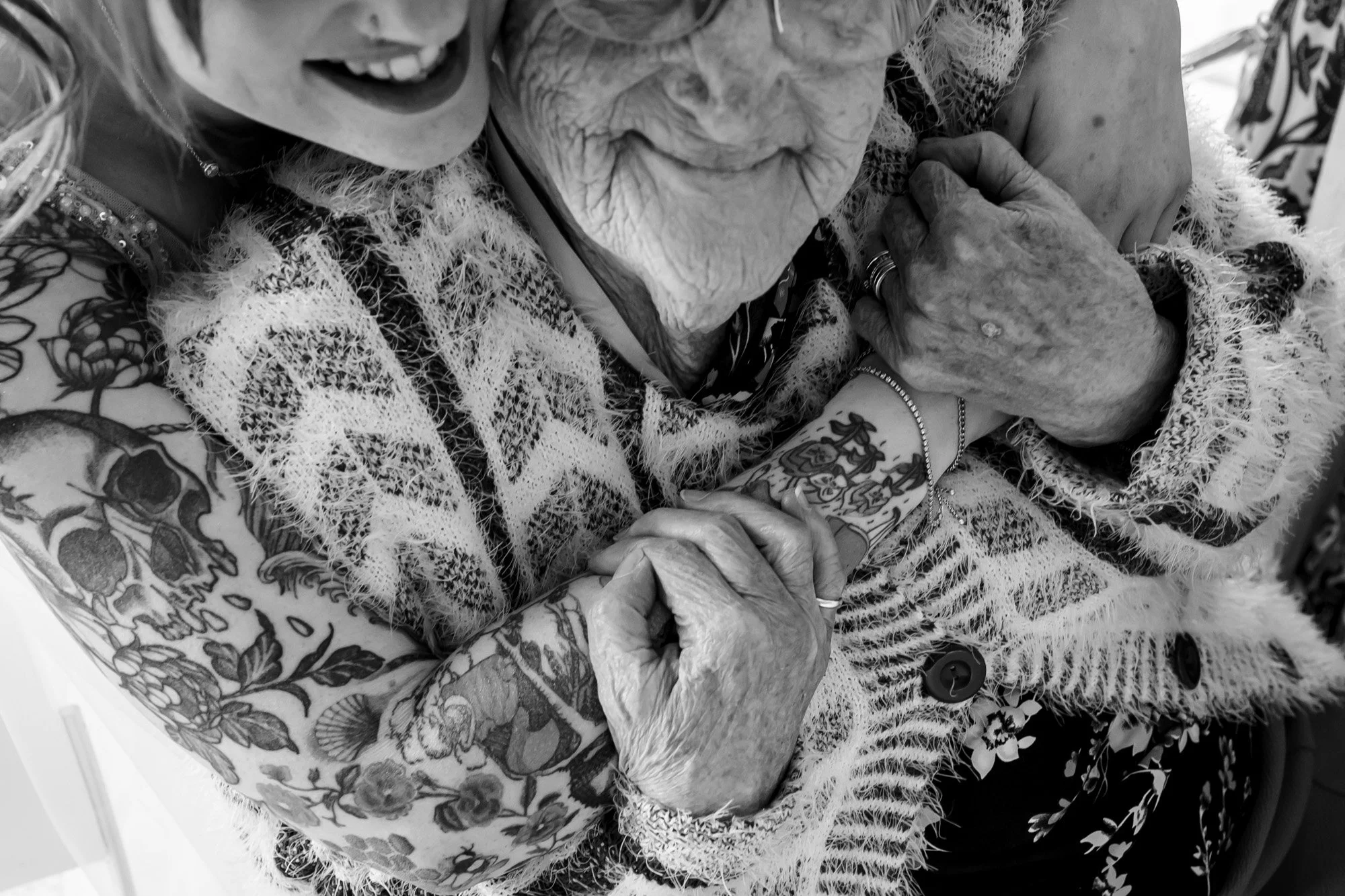 Close up black and white photograph of tattooed woman hugging her grandmother from behind. Their faces are not fully visible, but they are both smiling. The older woman is resting her hands on the younger woman's arm.