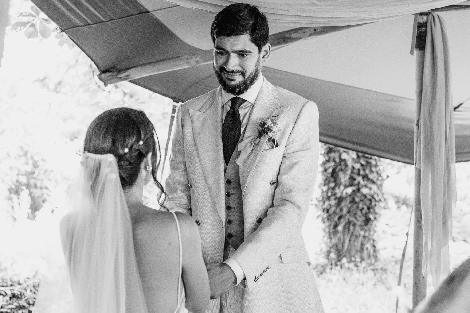 A bride and groom face each other during their wedding ceremony at tipis at whatton house. He looks emotional. Photograph by documentary wedding photographer Caroline Goosey