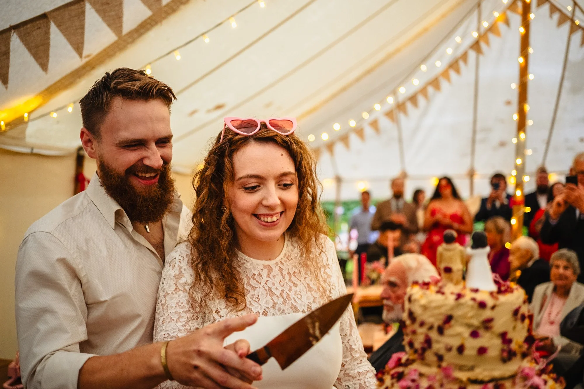 a young couple in casual wedding clothes are smiling as they both hold a knife to cut their cake. They're in a marquee decorated with festoon lights and bunting, and their guests can be seen watching in the background.
