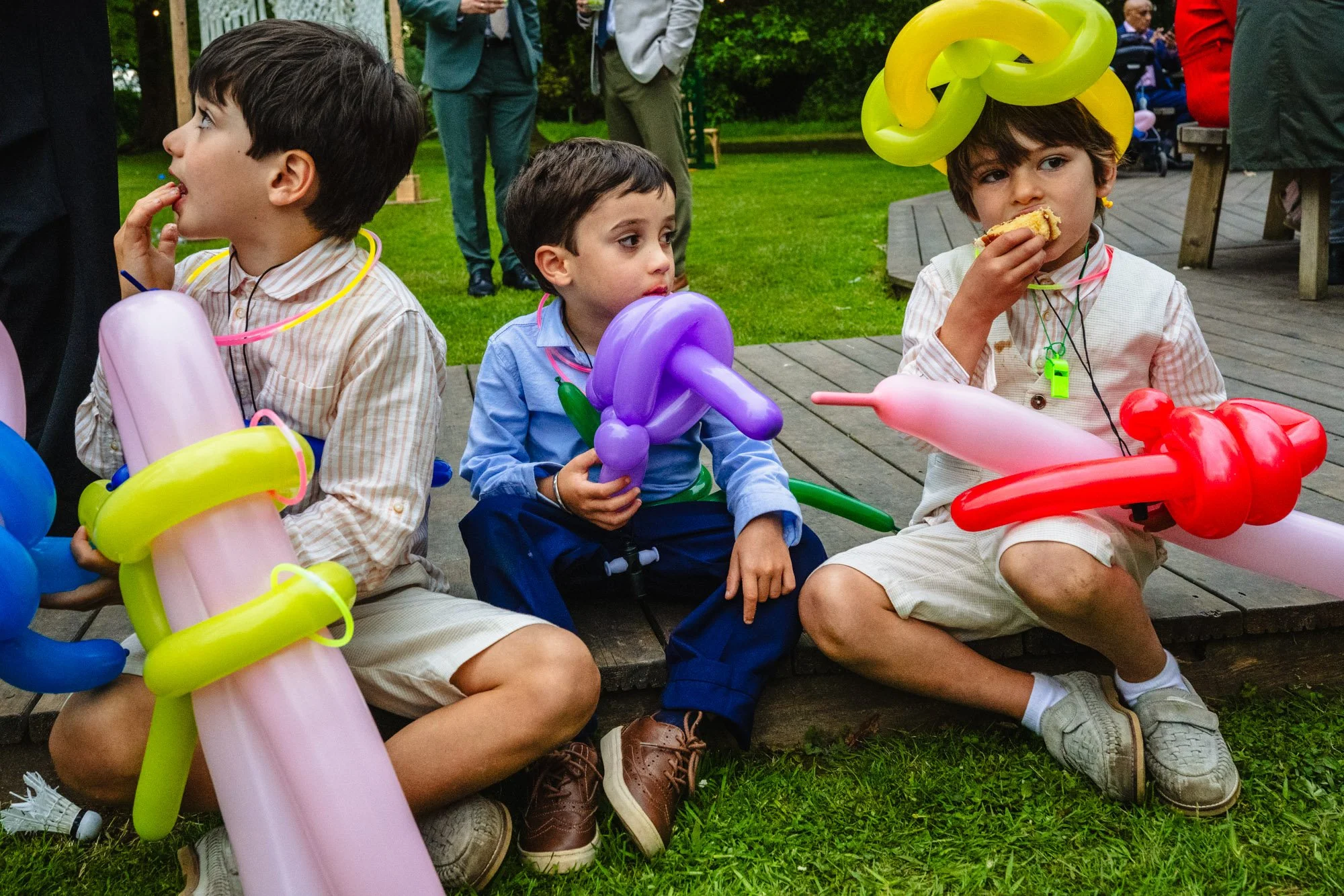 three young children with balloon guns sit on the deck at a wedding at whatton house tipis. The child in the middle looks tired, and the other two are eating cake.