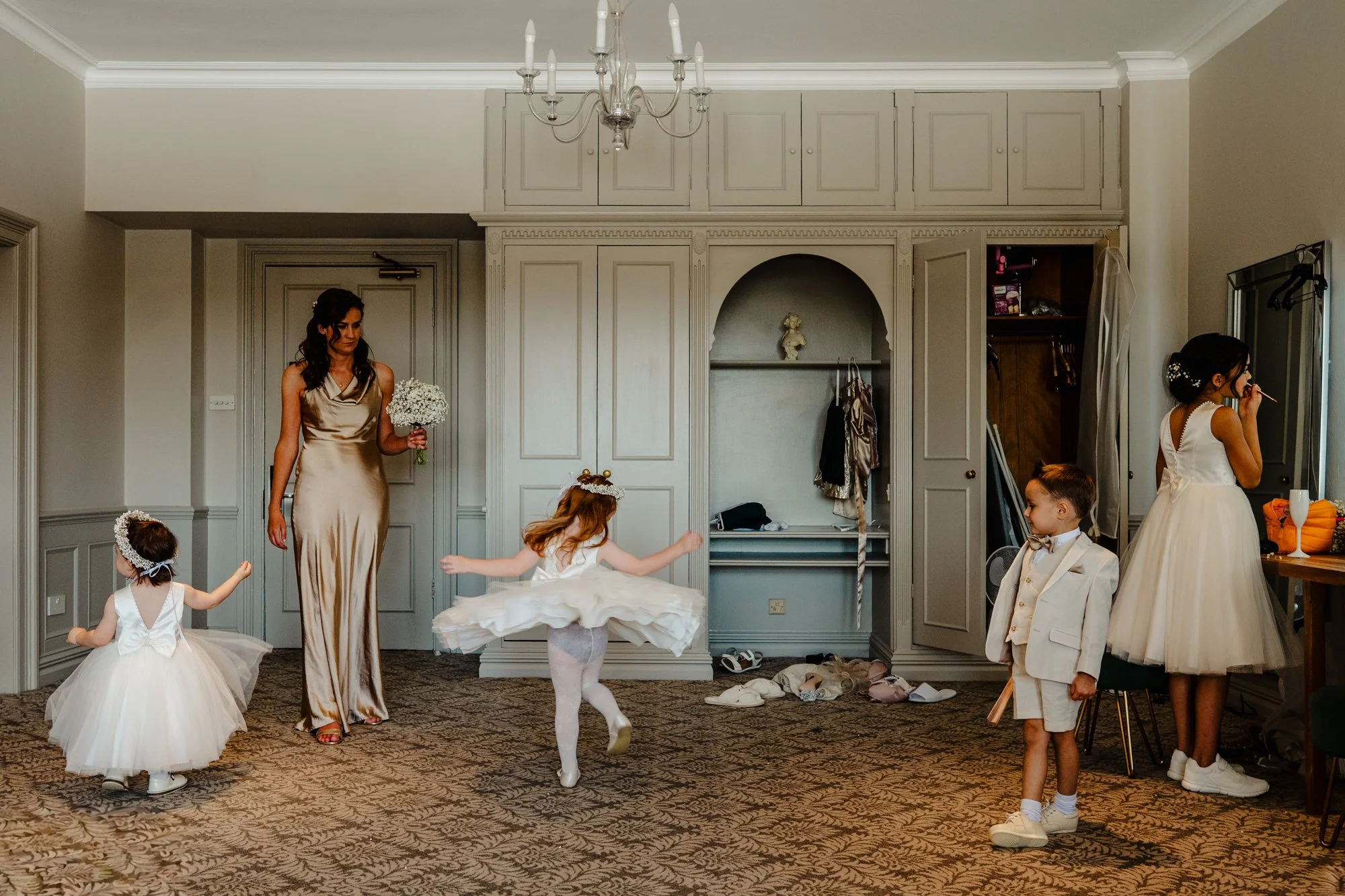 Documentary style wedding photograph taken in a large hotel room during the morning. Four children play, apply makeup and spin in circles while a bridesmaid watches on holding a bouquet of flowers