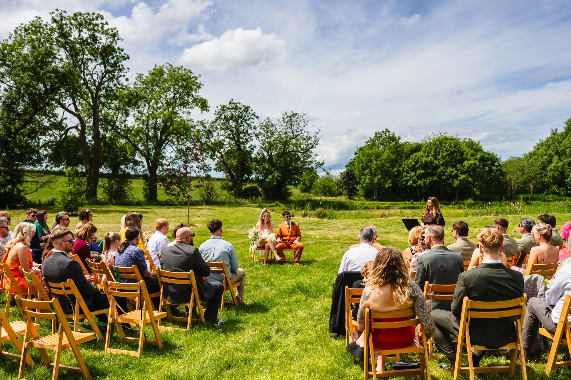 photograph of a wedding ceremony happening on the lawn at Duddon Mill Farm, Cheshire. The bride and groom sit on chairs while their guests are seated around them in a semicircle and a celebrant stands off to the right of the frame.