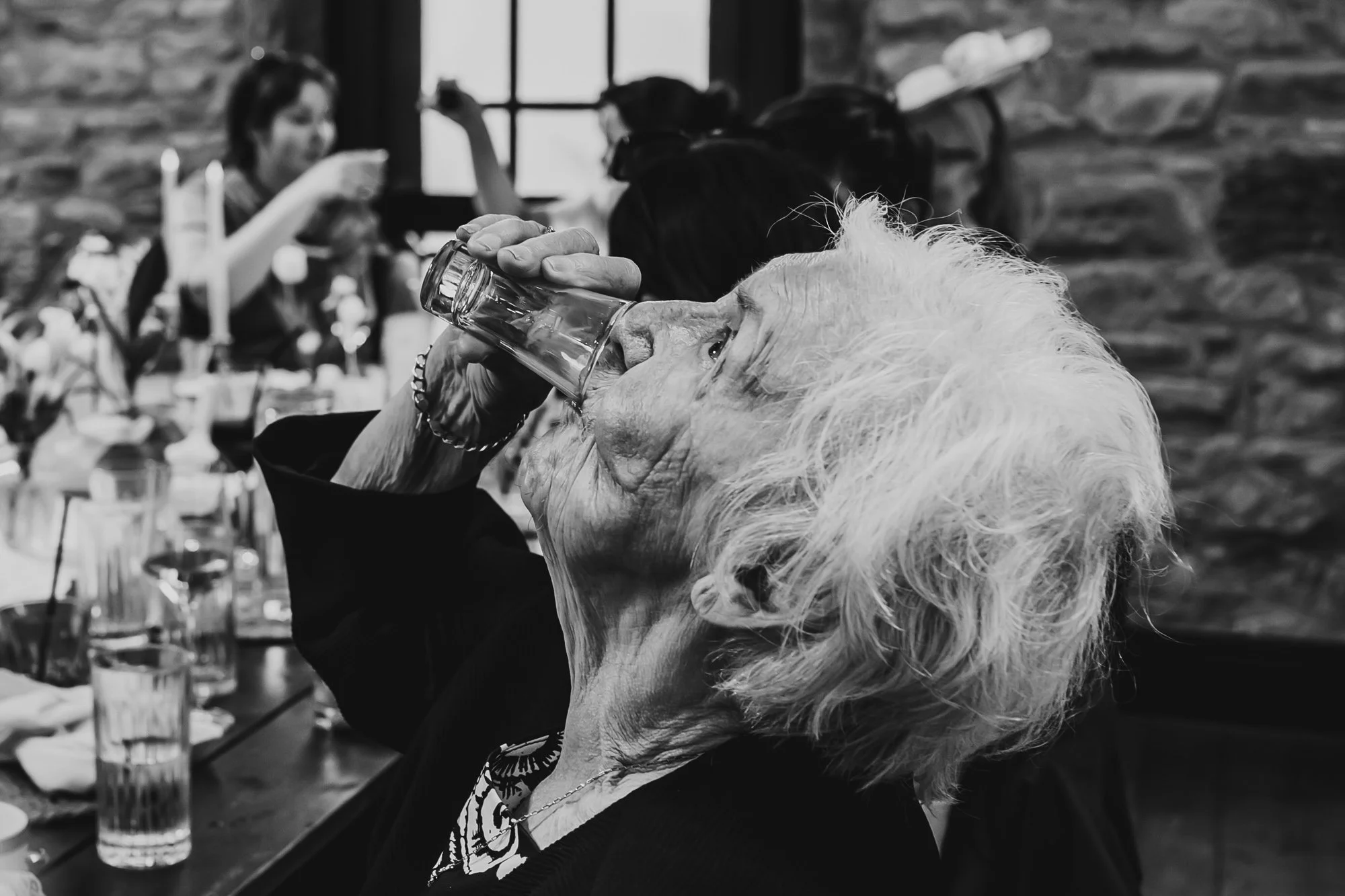 black and white photograph of an older woman drinking a shot of alcohol by documentary wedding photographer Caroline Goosey