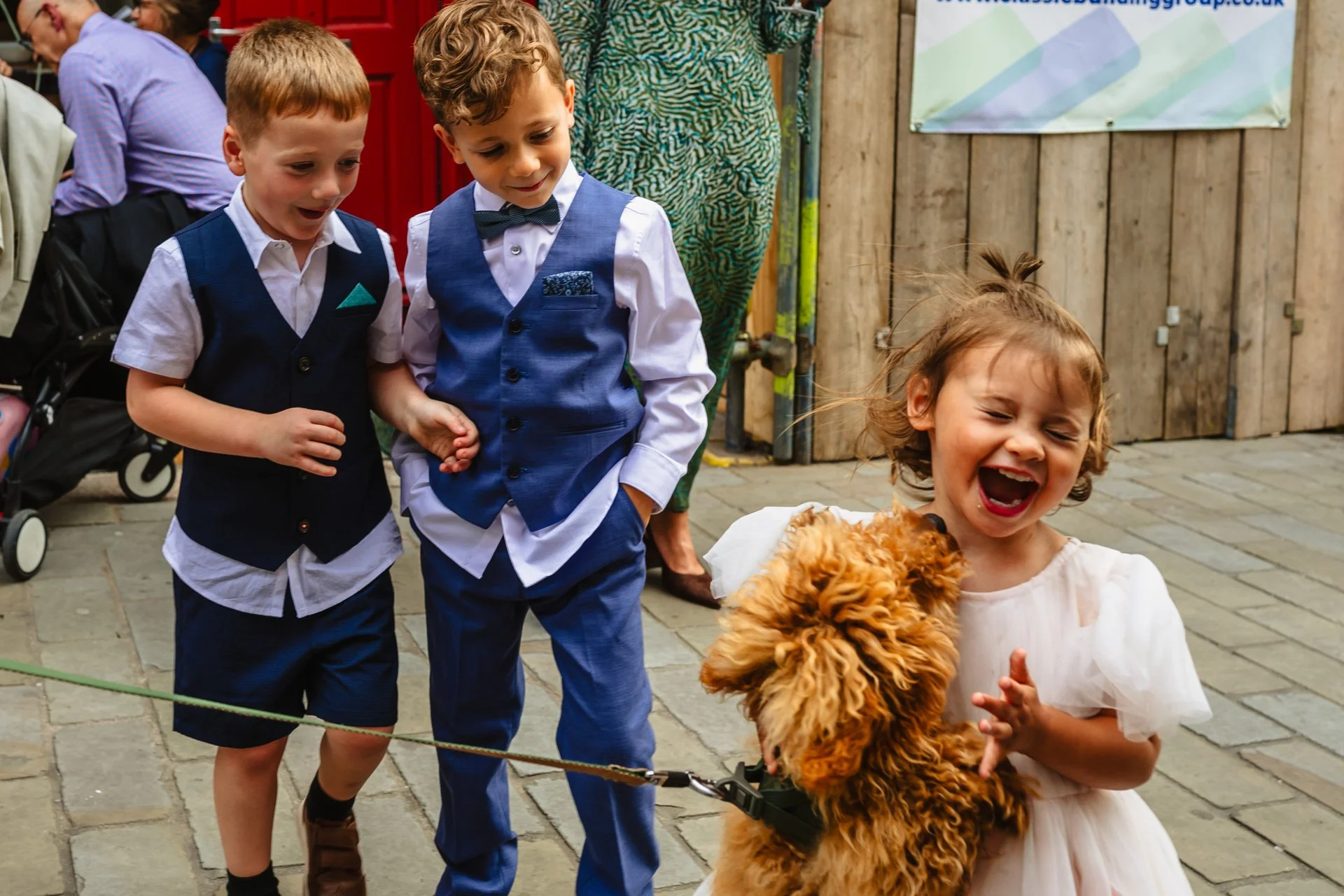 Three children and a small dog standing outdoors on a cobblestone surface. The children are dressed in formal attire, two boys in vests and one girl in a dress, all smiling and laughing, while the dog jumps up at the little girl.