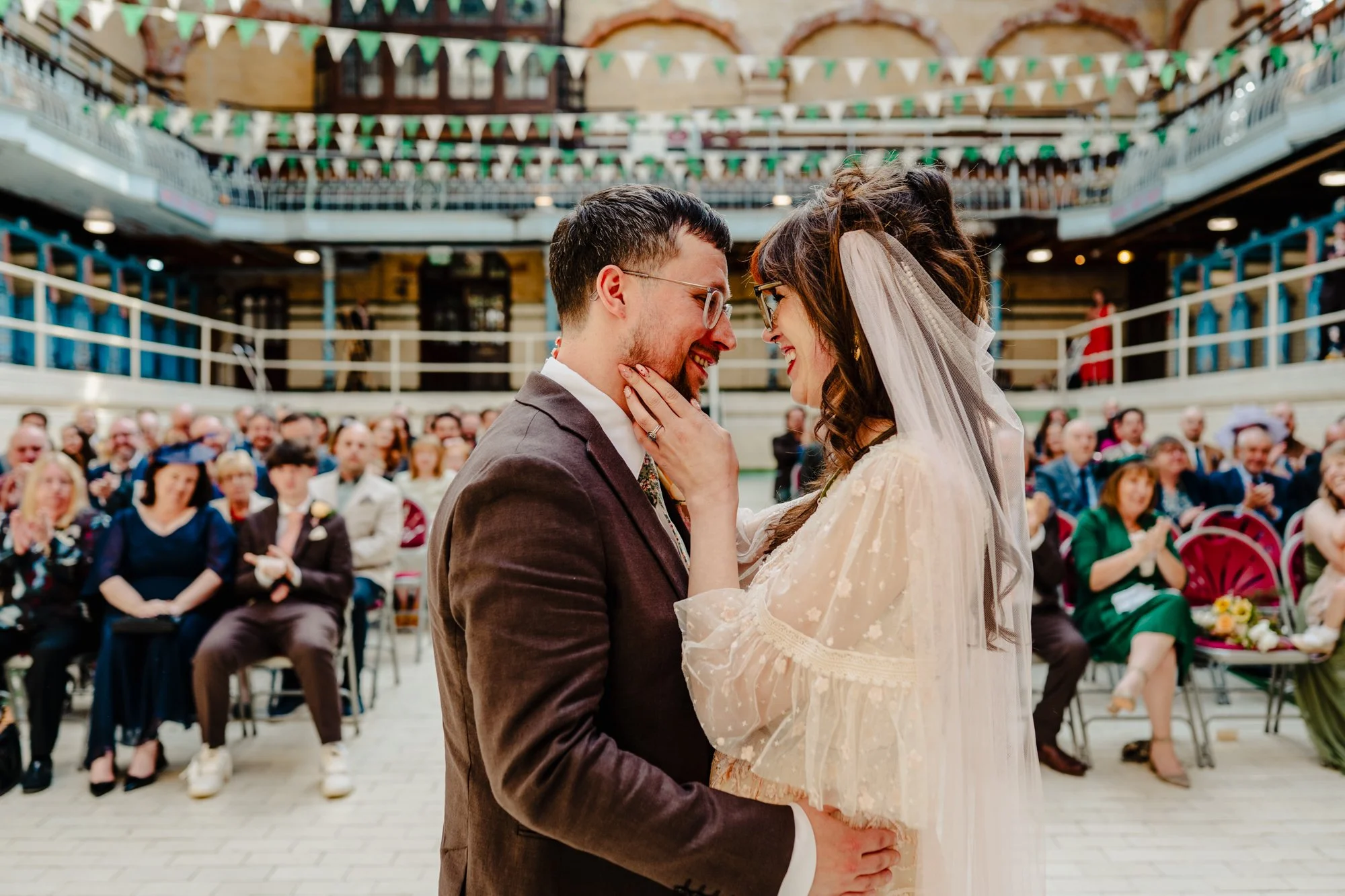 Photograph of a bride and groom embracing and smiling together during their wedding ceremony at Victoria Baths in Manchester.