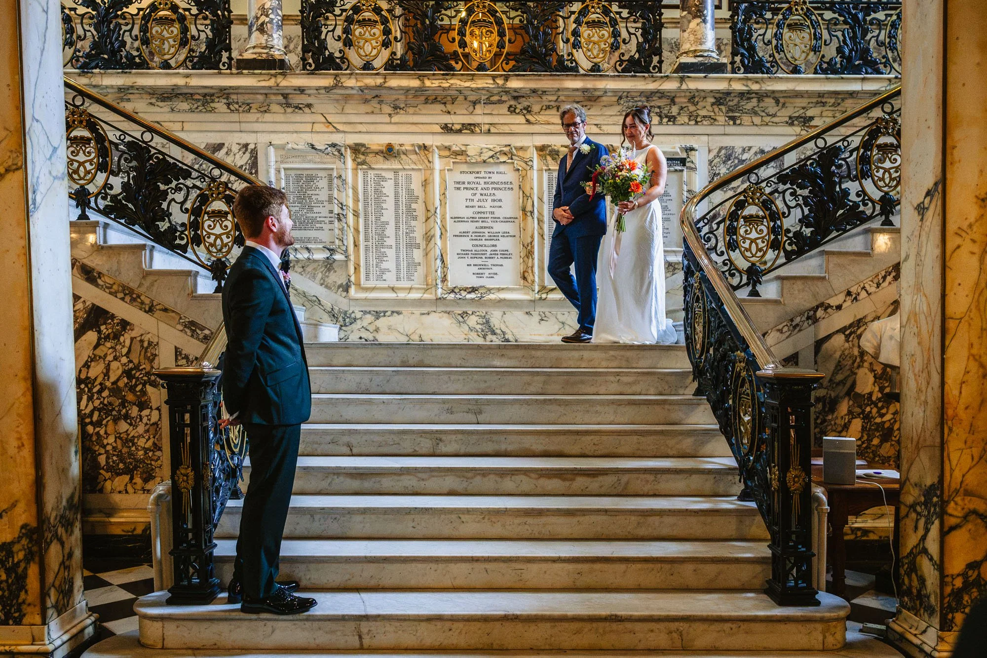 A bride descends the stairs at Stockport Town Hall with her father by her side. Her husband to be waits for her for their wedding ceremony to begin