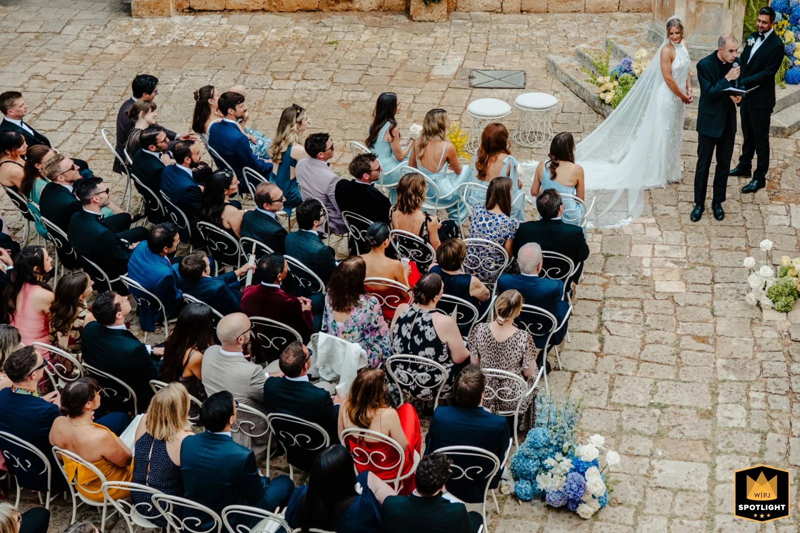 A wedding ceremony with a bride, groom, and officiant standing in front of seated guests on a cobblestone surface, with floral arrangements.