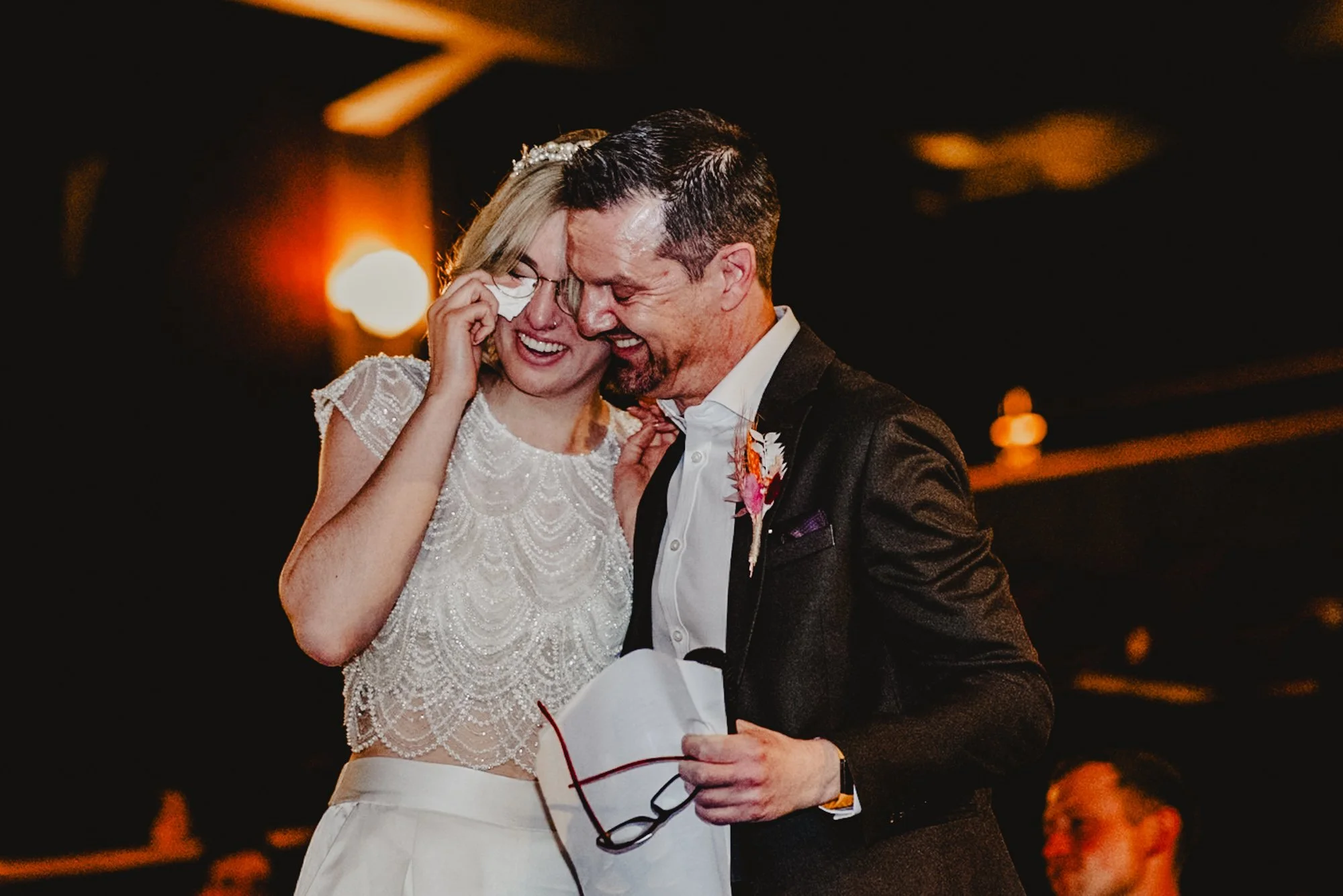 Photograph of a bride dabbing her eyes and smiling while she hugs her father at the end of the speeches at her wedding at Savoy Cinema in Heaton Moor, Manchester.