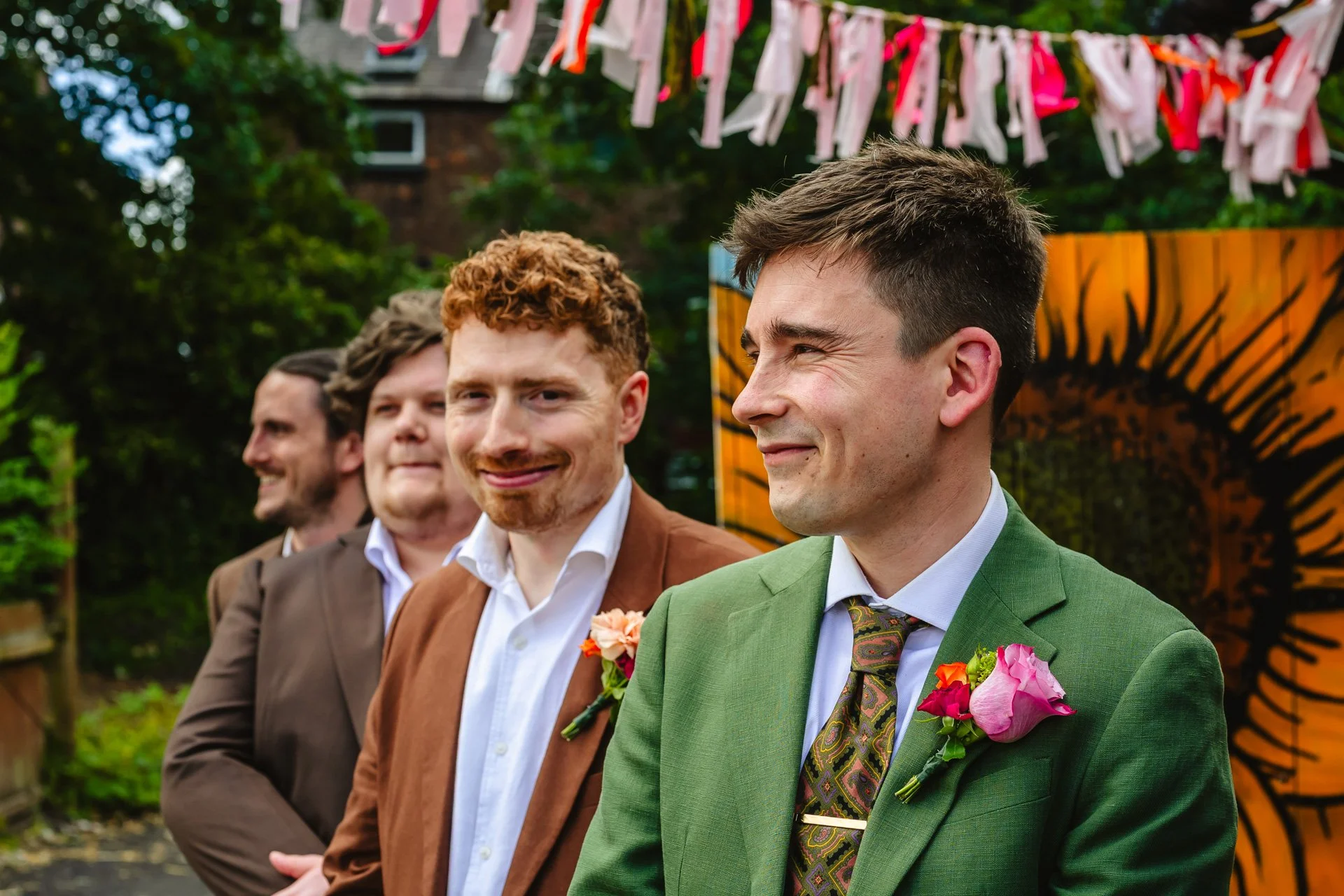 Four men wearing colorful suits and floral buttonholes at an outdoor event with decorative ribbons hanging overhead and vibrant orange and black art in the background.