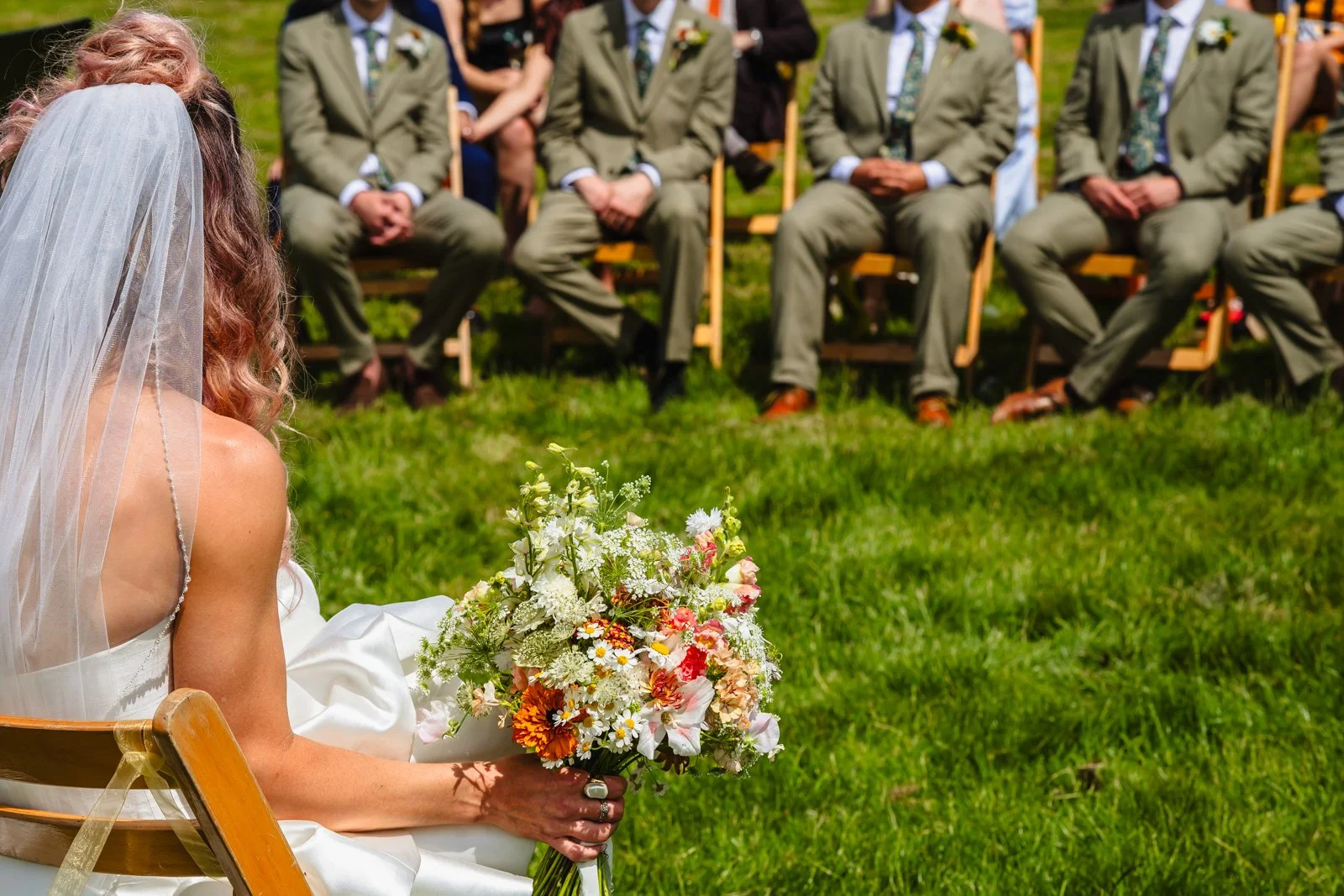 photograph of a bride holding a bouquet of wildflowers at an outdoor ceremony with the legs of her groomsmen visible in the background by Caroline Goosey Photography