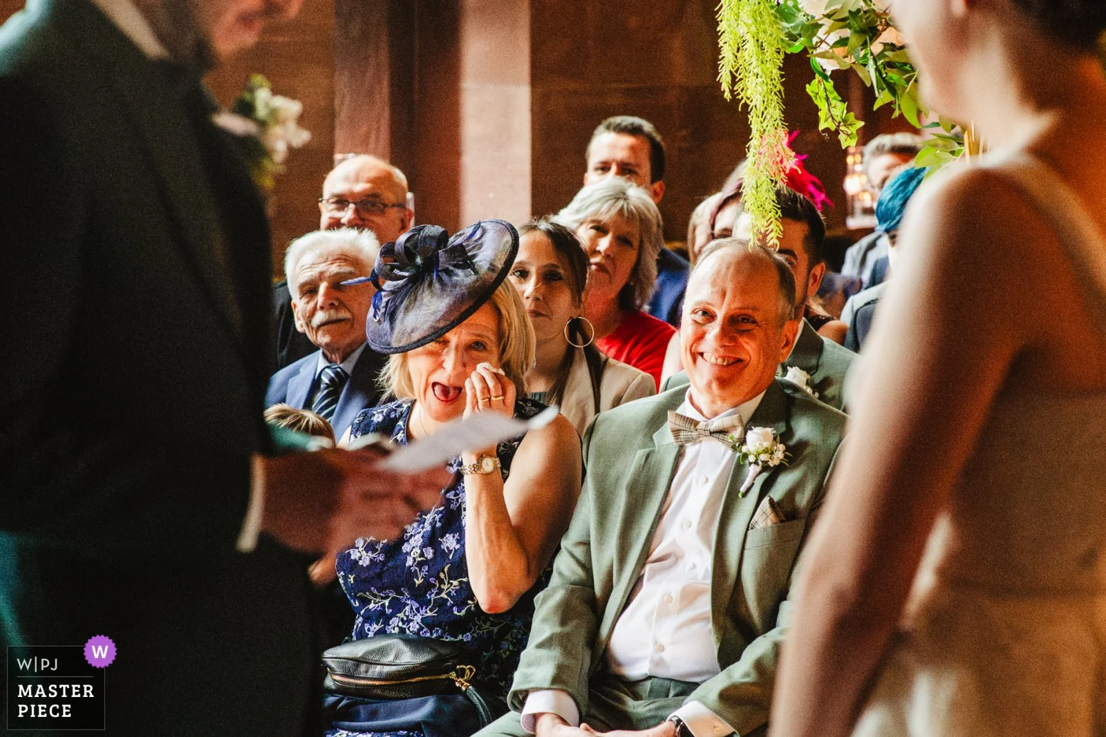 Photograph of a couple reading their vows to each other. Between them in the centre of the photo are the groom's parents looking emotional and happy. Taken at Peckforton Castle in Cheshire. An award from WIPJ is in the bottom corner of the image