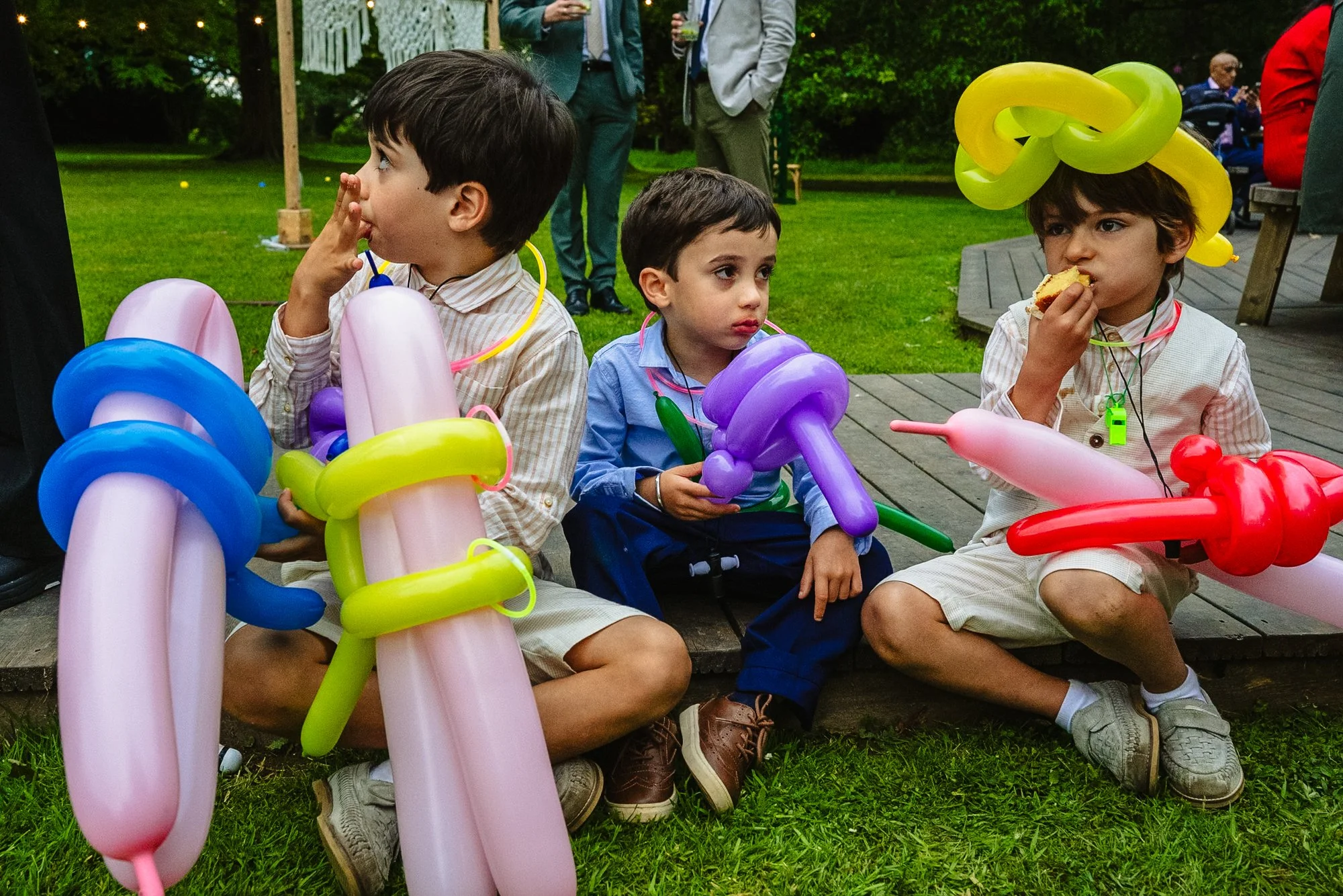 three young boys in dirtied formal clothing sit eating wedding cake and looking tired. They have balloons in the shapes of hats and guns, rave whistles and glow sticks.