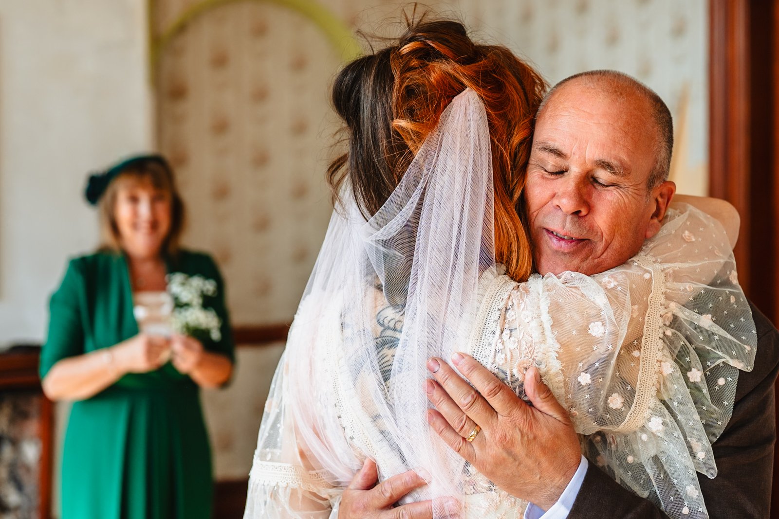 photograph of bride hugging her father while her mother watches in the background, her smile just about visible at Victoria Baths in Manchester