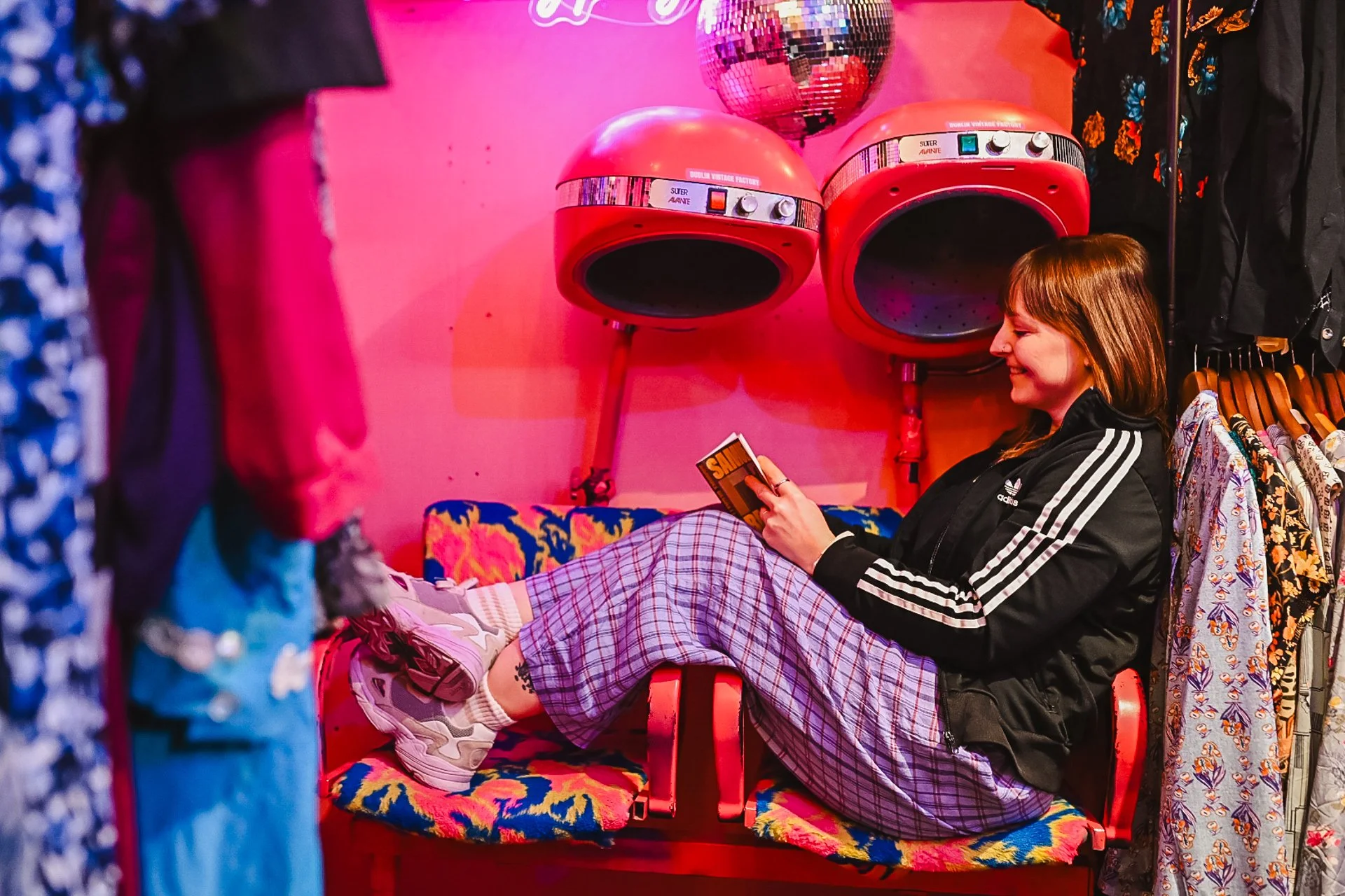 Photograph of a woman wearing purple checked trousers and an adidas zip top. She is sitting sideways on a chair in a pink room.