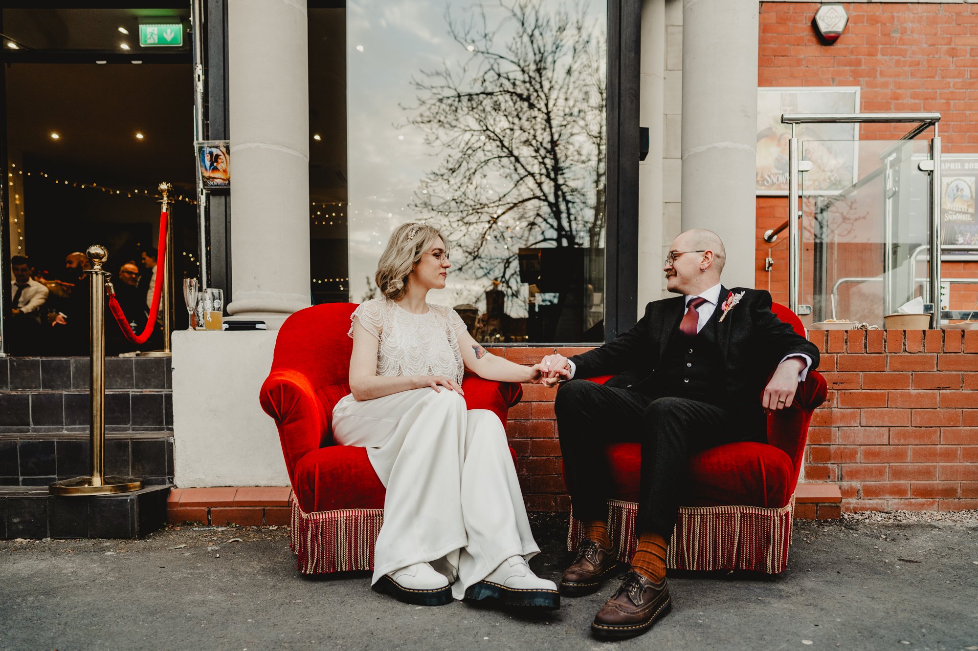 Wedding photography of a bride and groom sit on red velvet chairs on the pavement outside the Savoy cinema in Heaton Moor. She is wearing trousers and a beaded ivory top, he is wearing a suit. They are both wearing Dr Martens.