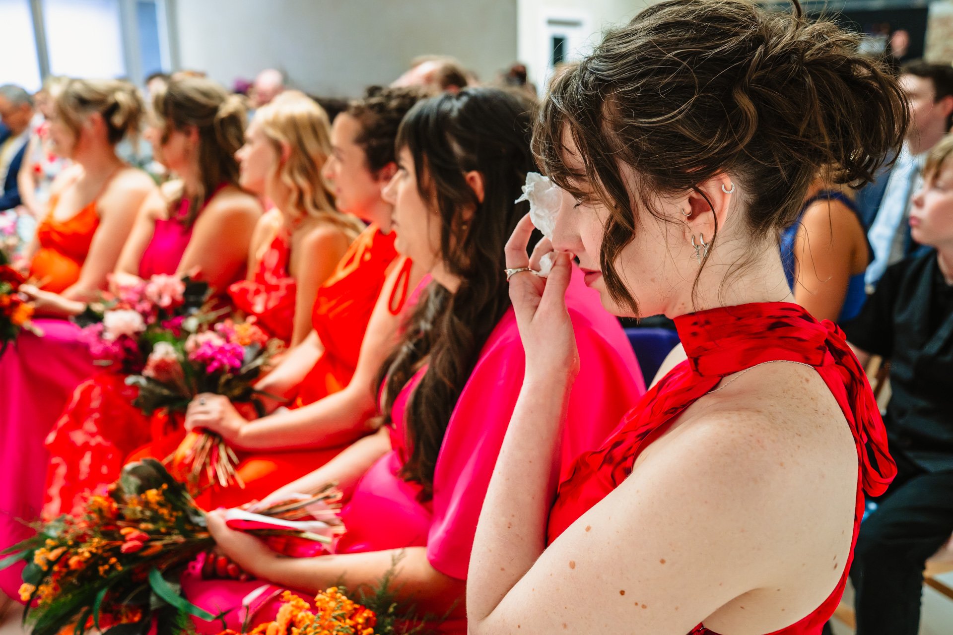 A group of women dressed in red and pink dresses, sitting with flowers on their laps, at a wedding ceremony in Liverpool.