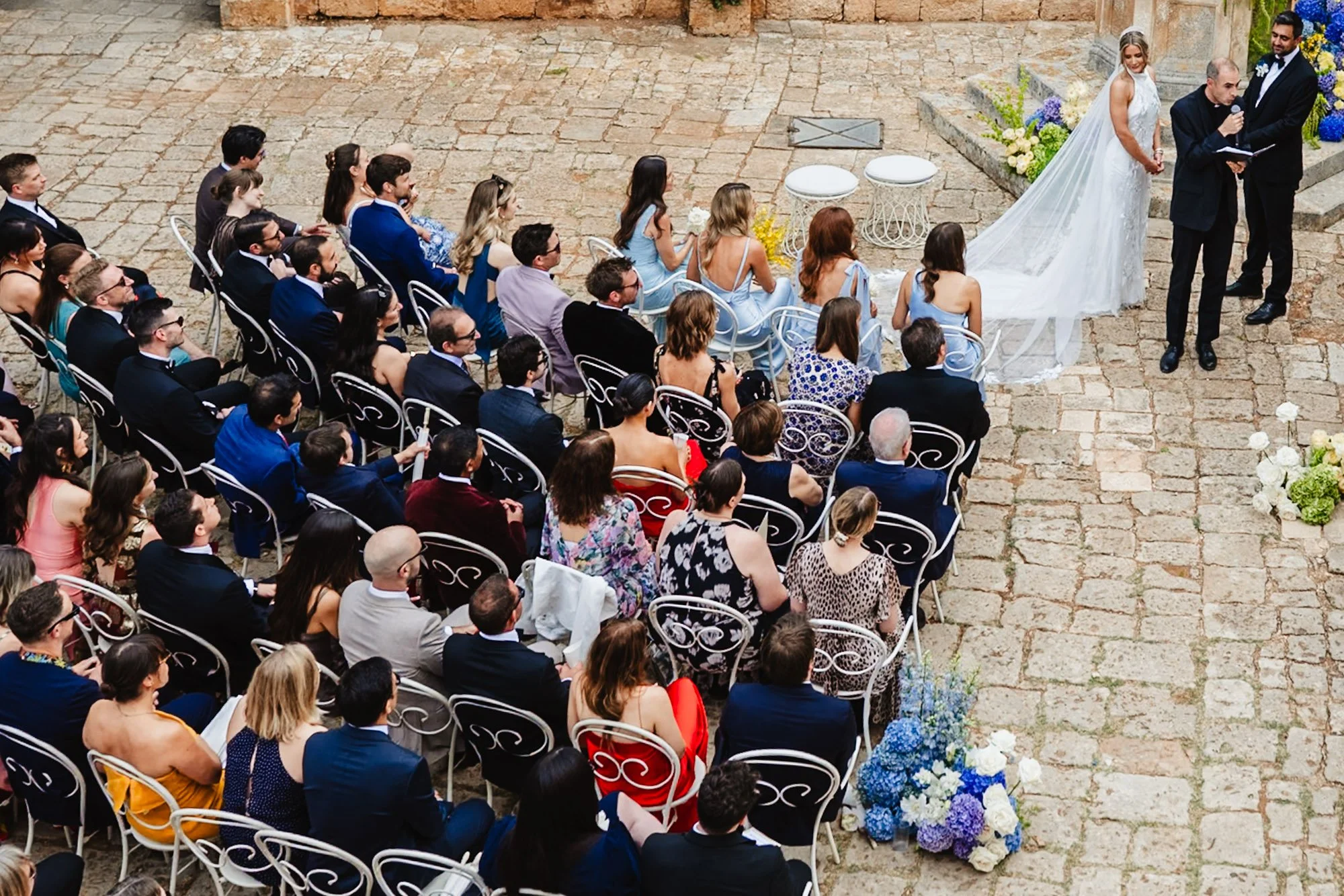 A photograph of an outdoor wedding ceremony in full flow, taken from a high elevation. The couple are in the left top corner with their vicar, while the guests occupy three quarters of the rest of the space. Blue and purple hydrangea decorate the ais
