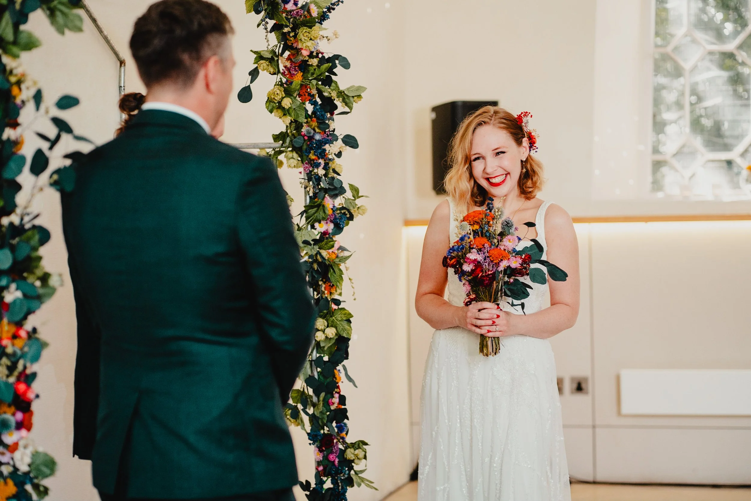 A bride in a white lace wedding dress holding a bouquet of colorful flowers, smiling at a groom during a wedding ceremony, with a floral arch in the background.