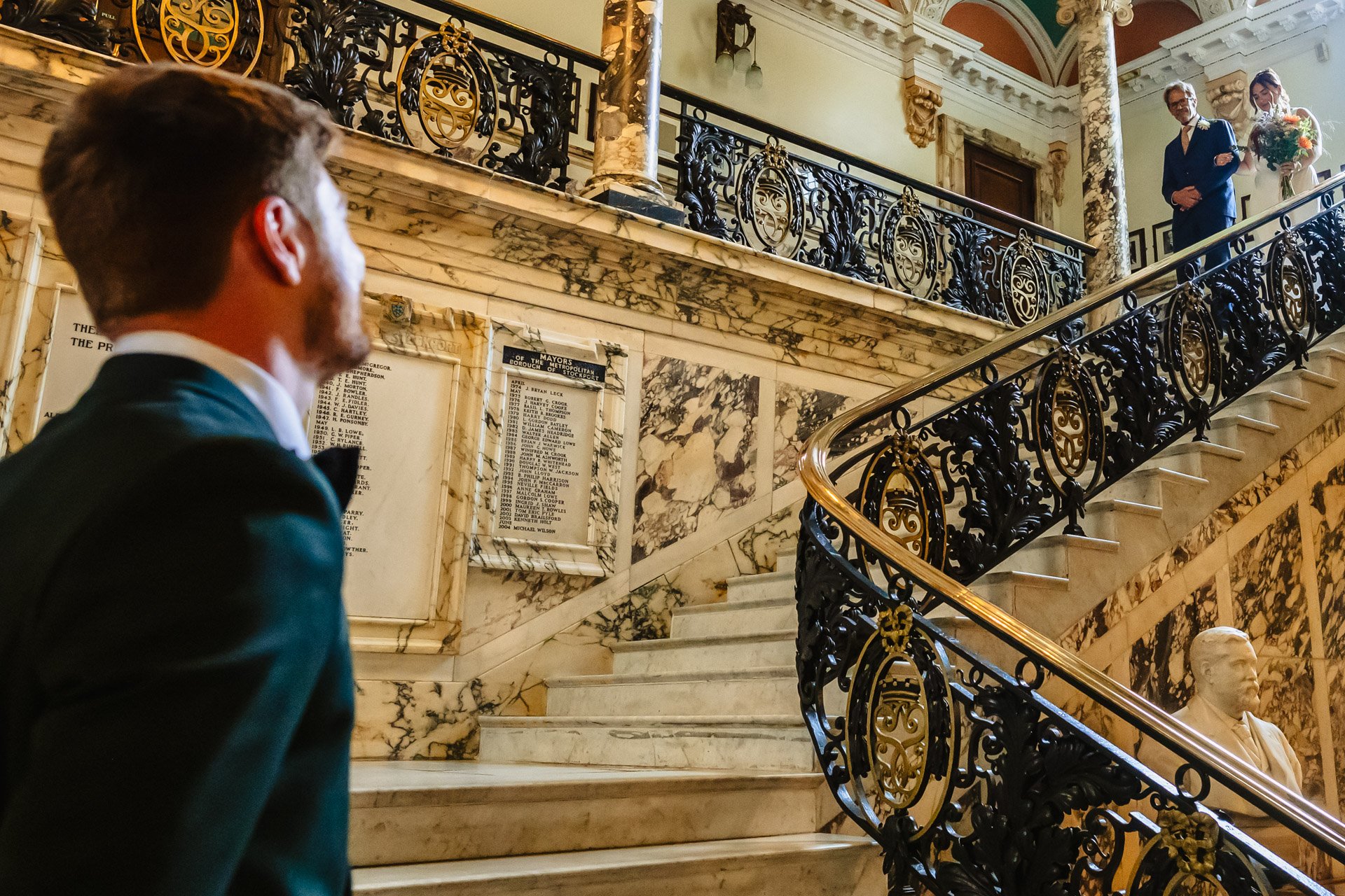 A man in a tuxedo is standing on the marble staircase at Stockport Town Hall. There is a woman in a white dress at the top of the staircase and a man in a tuxedo. 