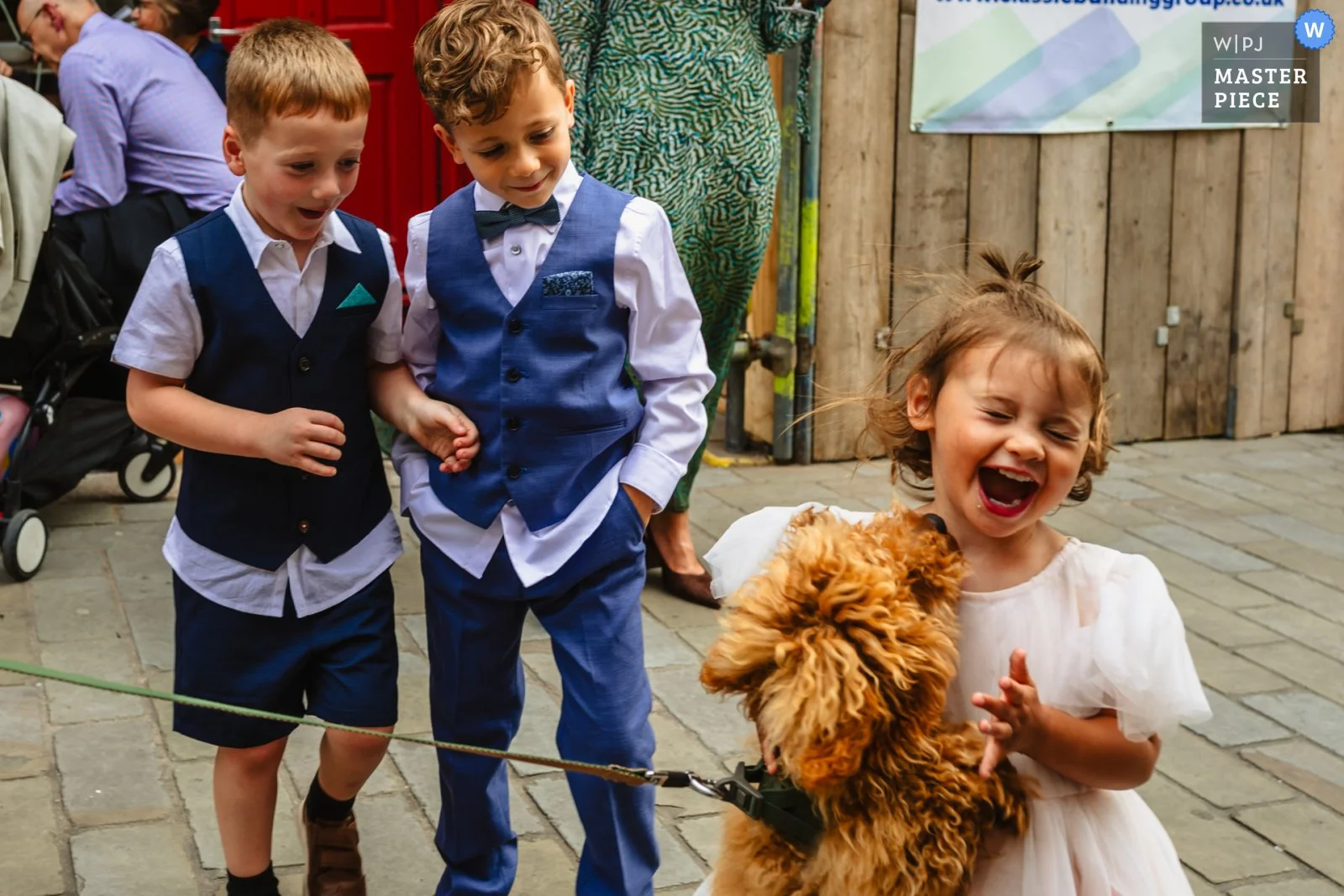 A young girl in a light pink dress laughing and holding a fluffy brown puppy on a leash, with two boys in formal attire looking at her, all outdoors on a stone-paved area.