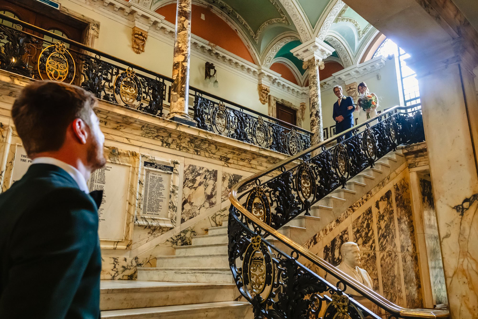 Photograph of a bride entering her ceremony on the marble staircase at Stockport Town Hall while her groom waits at the bottom