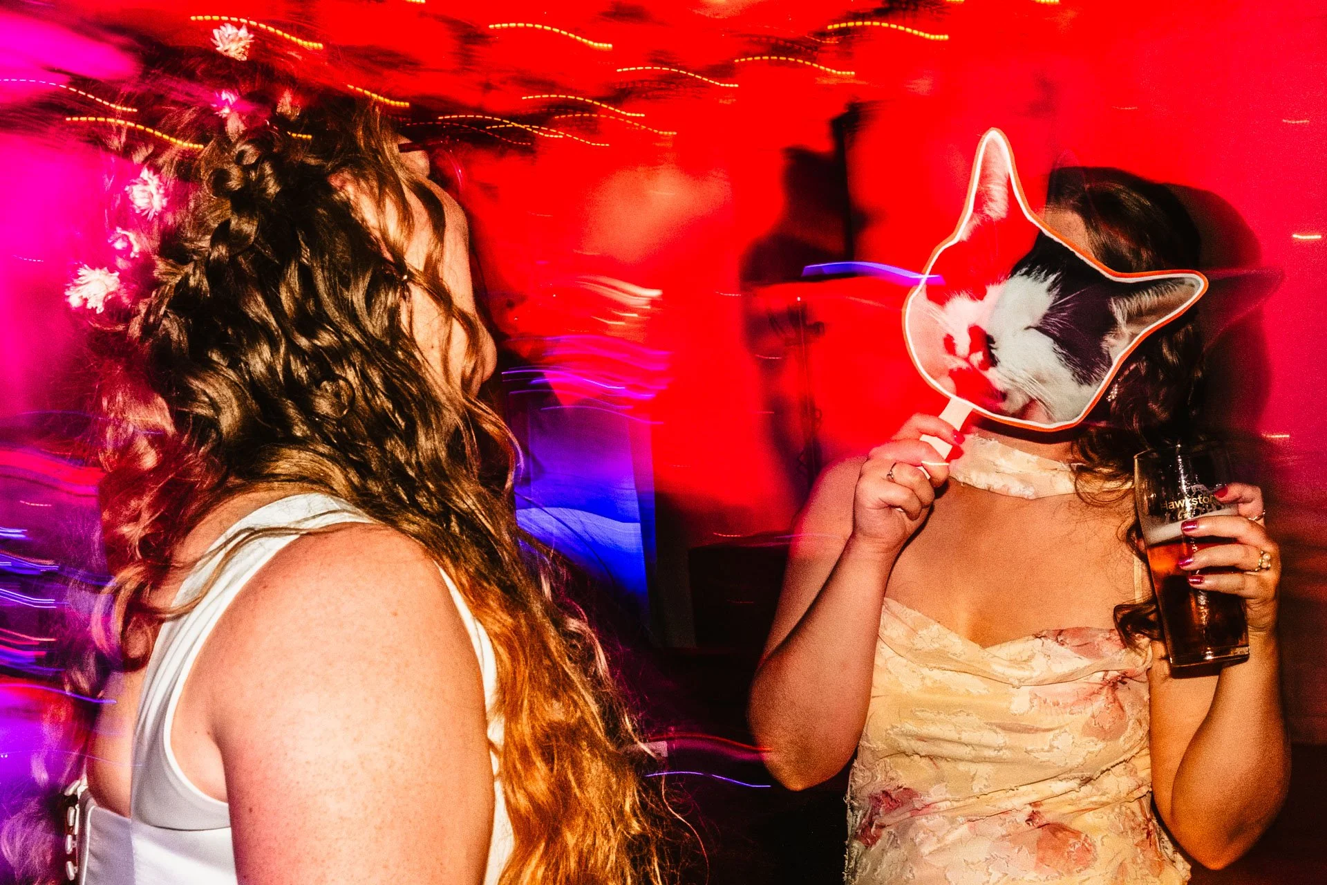 photograph or two women dancing at a wedding. One is holding a cut out mask of a cat's face in front of her own face.