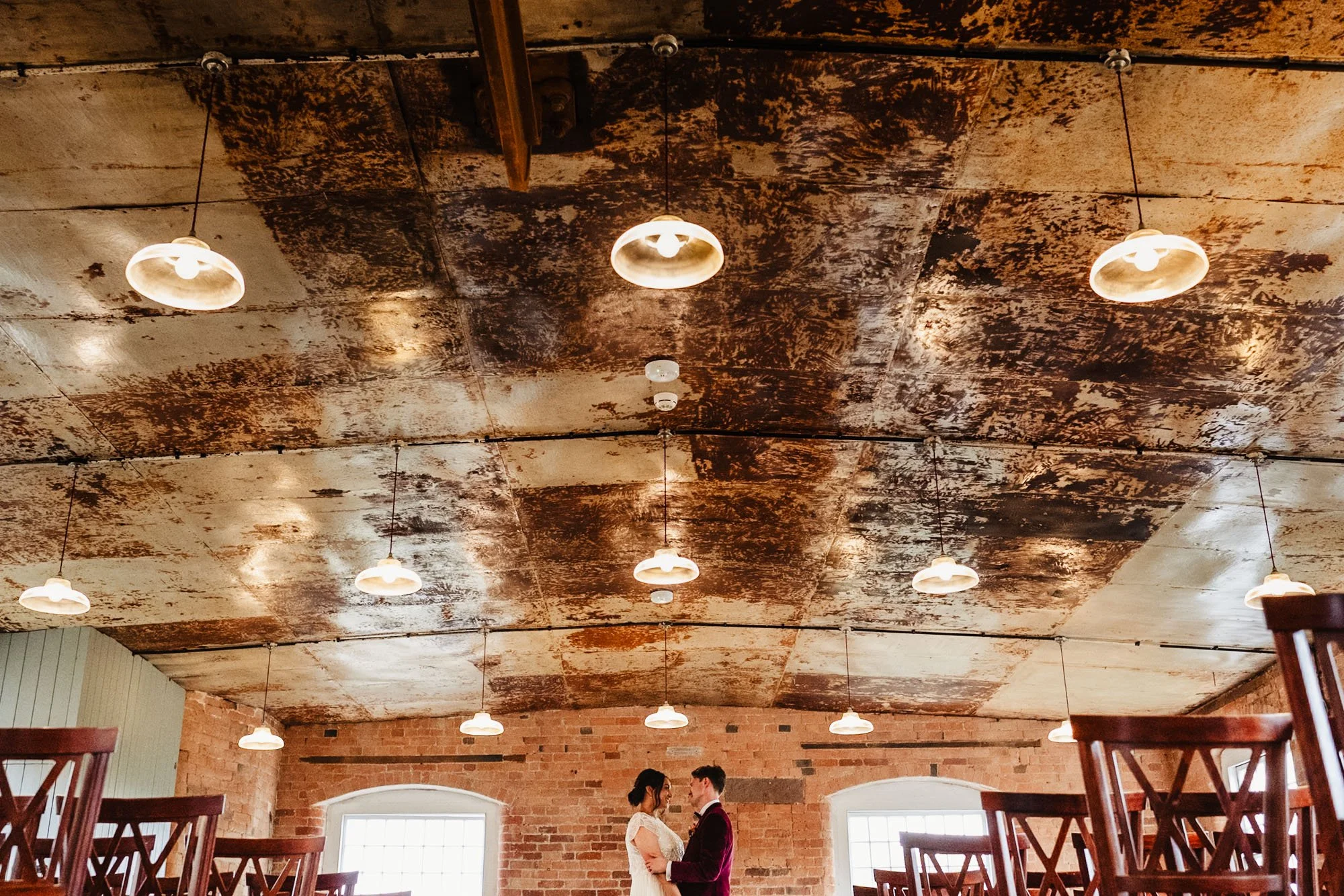 Wide angle photograph of a couple sharing a quiet moment at their wedding at West Mill in Derby. The ceiling is lit with many pendant lights.