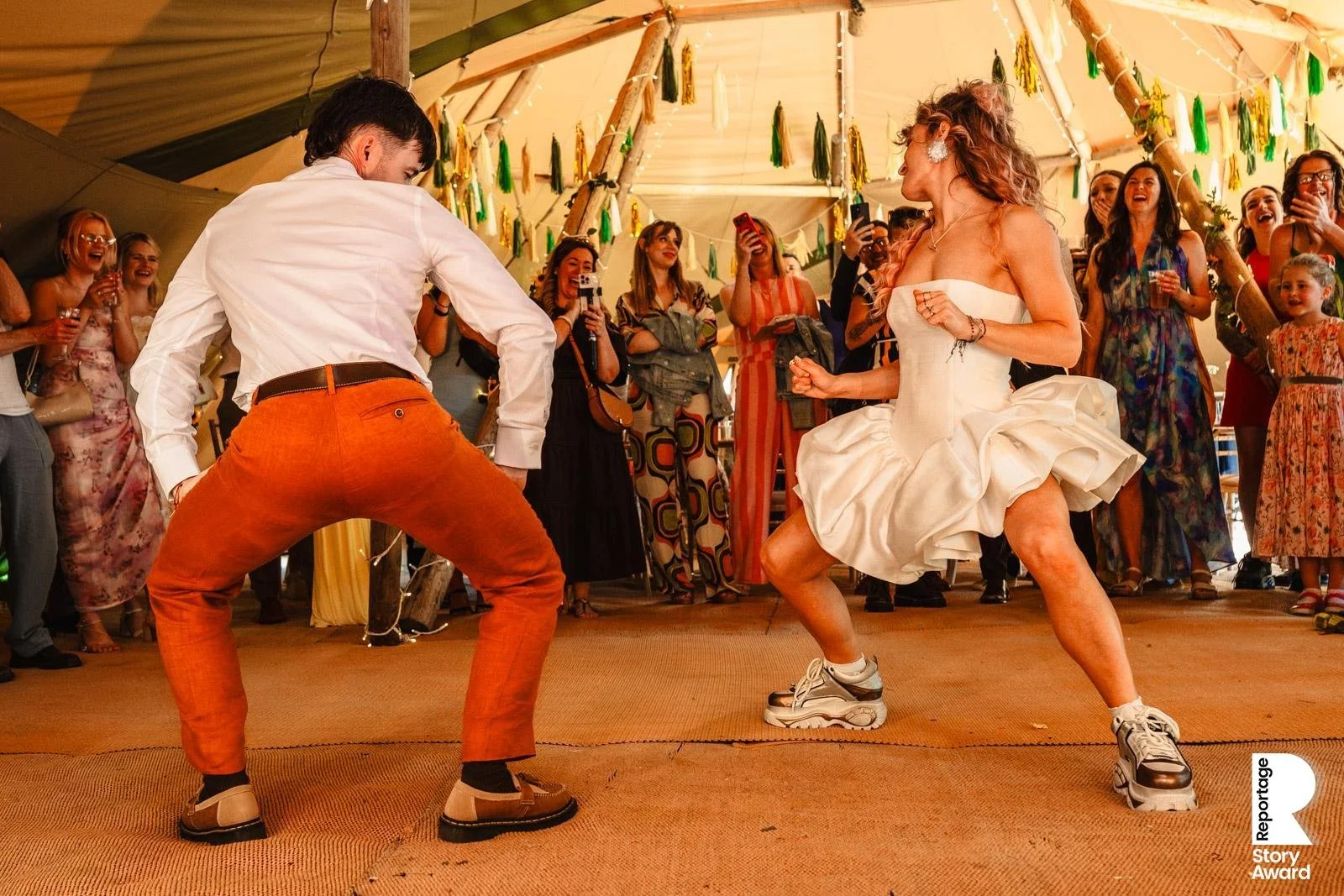 A lively scene at a wedding reception showing a woman in a white dress and sneakers dancing with a man in orange pants and a white shirt. They are surrounded by guests watching and smiling, under a decorated tent with hanging green and yellow streame
