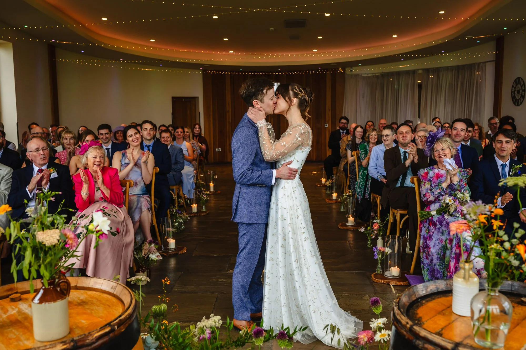 photograph of a bride and groom having their first kiss while all their guests smile and clap at The Out Barn in Clitheroe, by natural wedding photographer Caroline Goosey