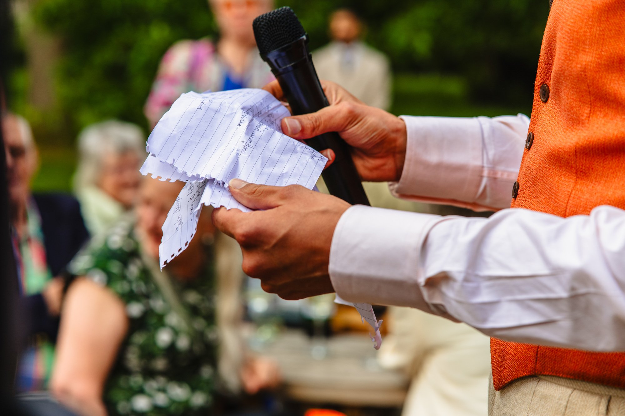 unpose photograph of a groom in an orange waistcoat holding a handwritten speech on a scrap of paper during his wedding speech by documentary wedding photographer Caroline Goosey
