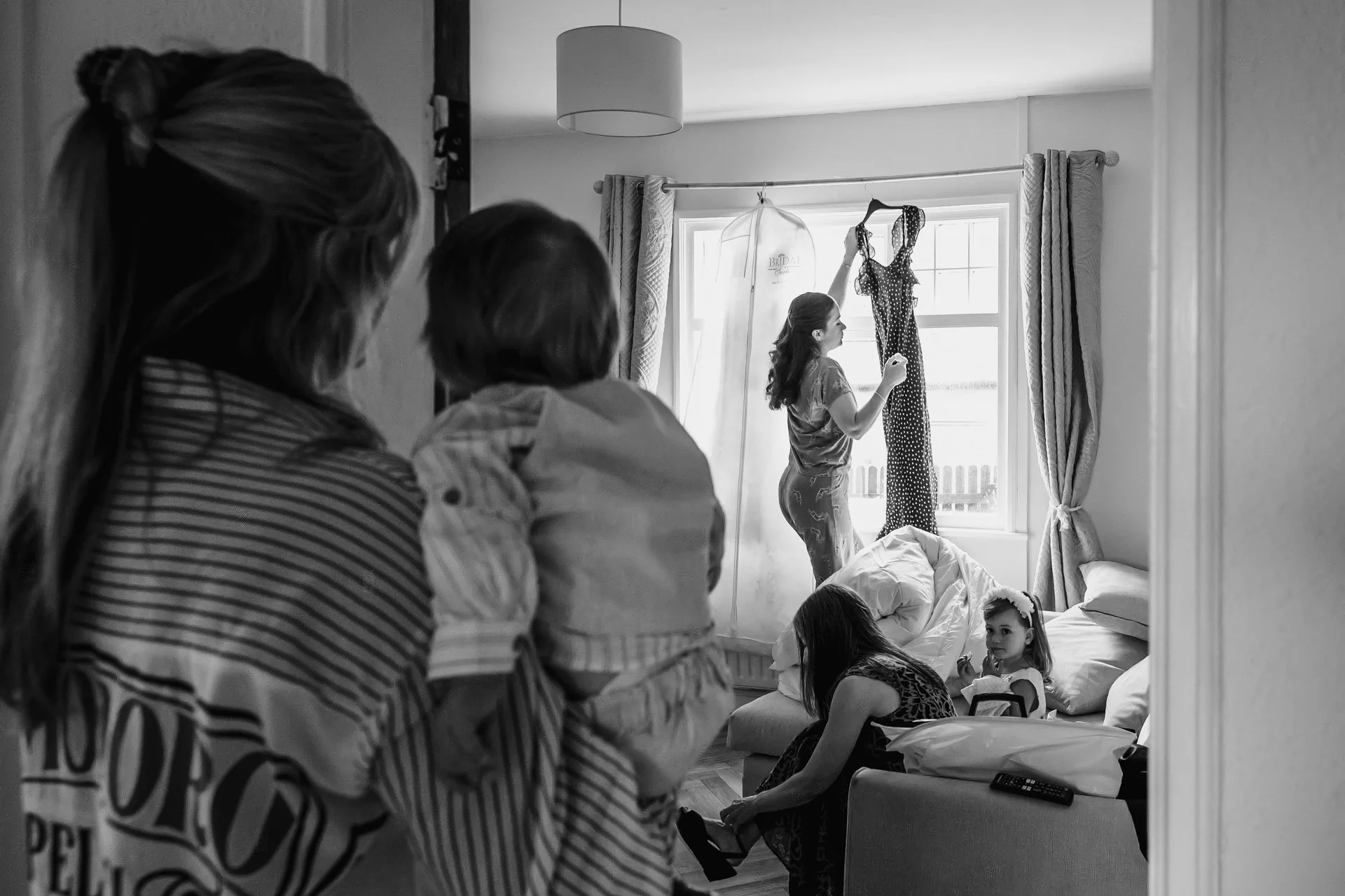 A group of women and children gathered in a bedroom, with some standing and some sitting on the bed. One woman is hanging a polka dot dress on a hanger by the window, and another woman is holding a baby. The room has curtains, a ceiling light, and a 