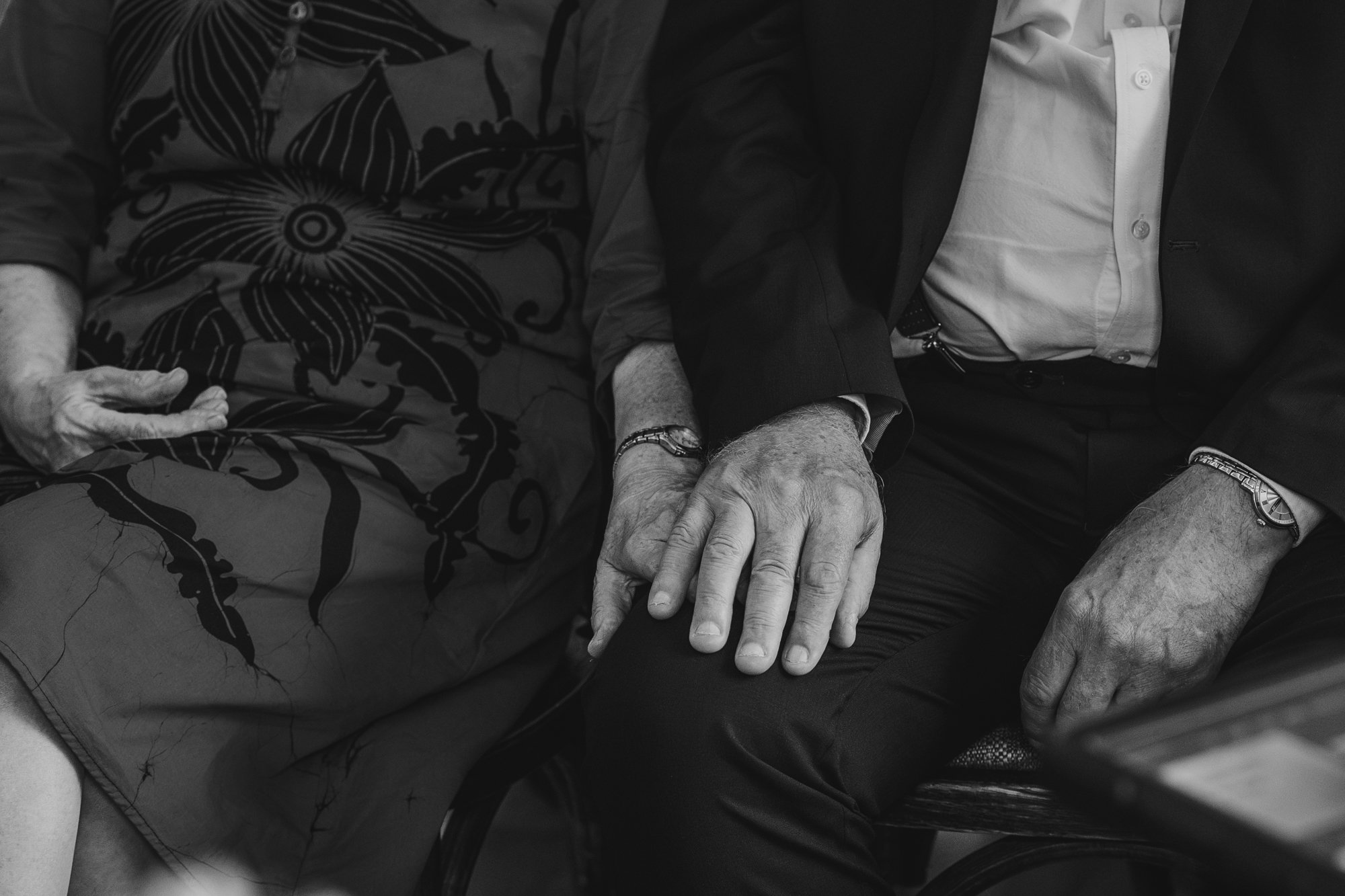 Close-up image of two men holding hands on their laps, seated next to each other. One man is wearing a patterned shirt and the other is in a suit jacket and white shirt.