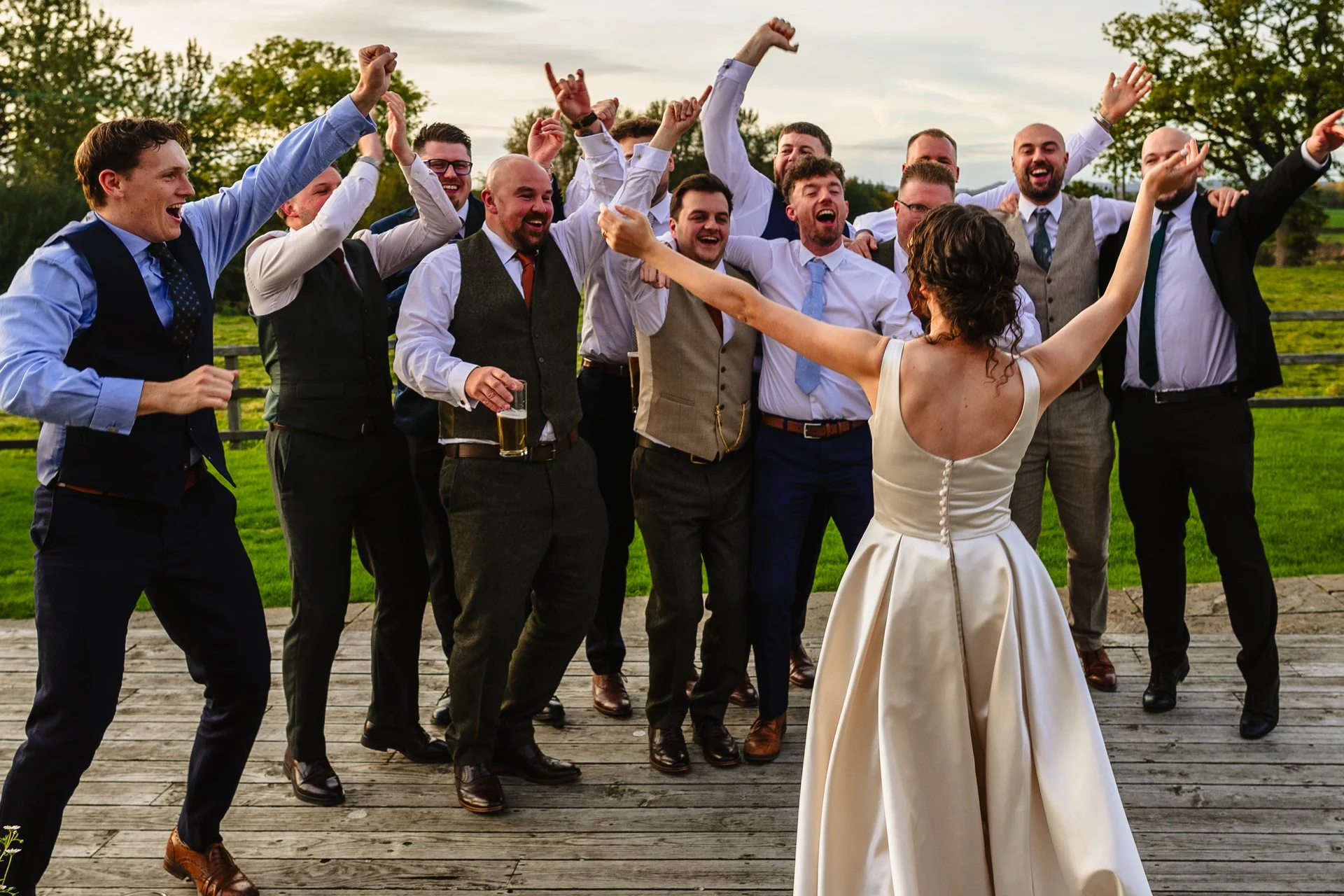 a group of men in smart clothing cheer in front of a woman as she stands facing them, arms raised. They are at a wedding at Grange Barn in Cheshire.