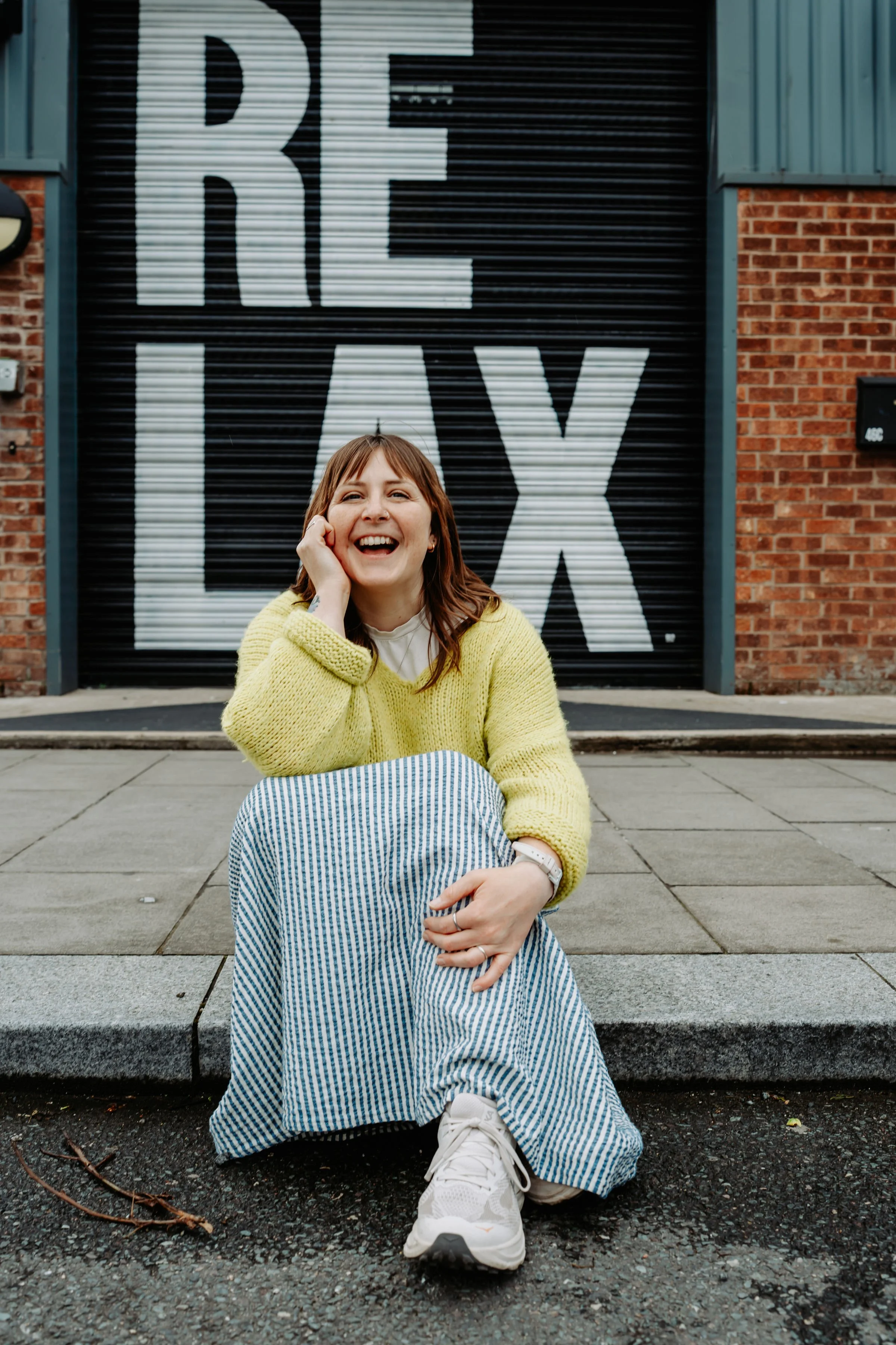 A young woman sitting on steps in front of a large black and white sign that says 'RE LAX'. She is smiling, wearing a yellow sweater, striped wide-leg pants, and white sneakers, with her hand resting on her face.
