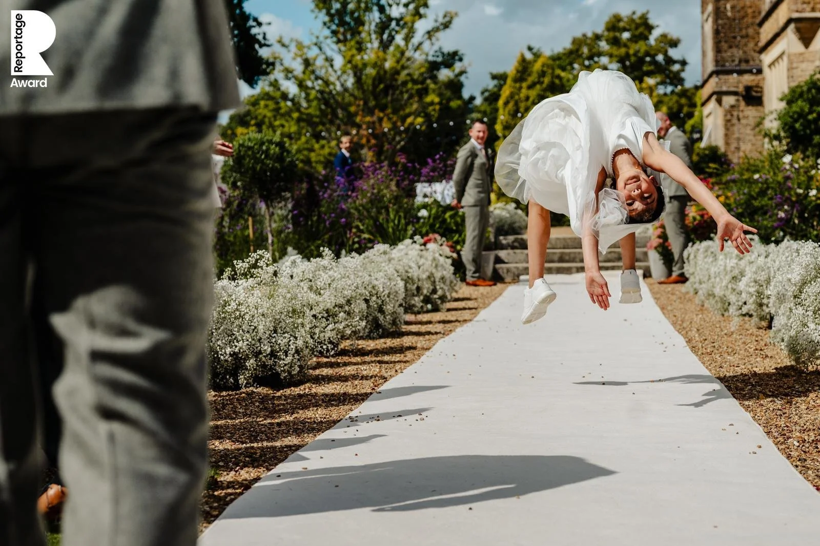 Photograph of a bridesmaid backflipping down the aisle. This photo has won multiple wedding photography awards, and has one from This Is Reportage displayed in the corner.