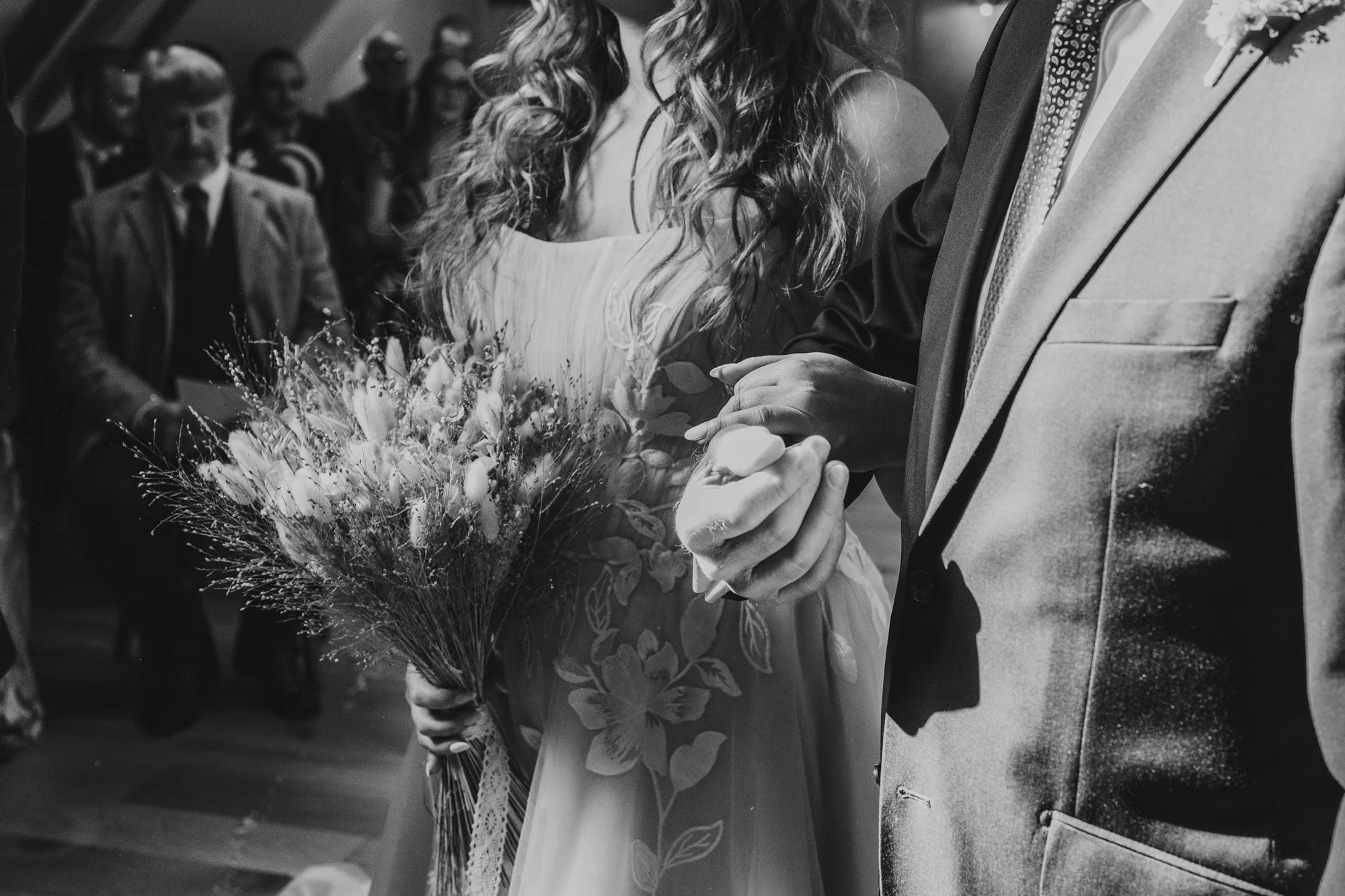 Black and white close up image of a bride holding a bouquet in one hand and her father's arm in the other as she walks down the aisle at Moat Hall Barns in Shrewsbury