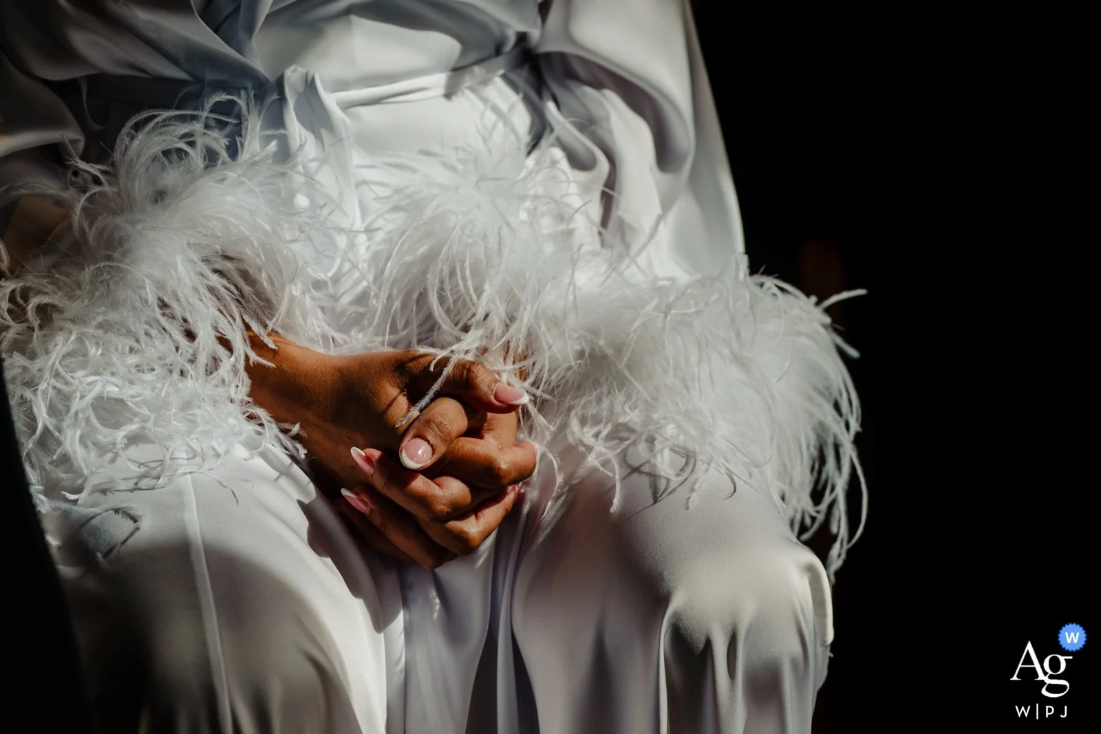 A person dressed in white with feathered details on their clothing, sitting with hands clasped, against a dark background.