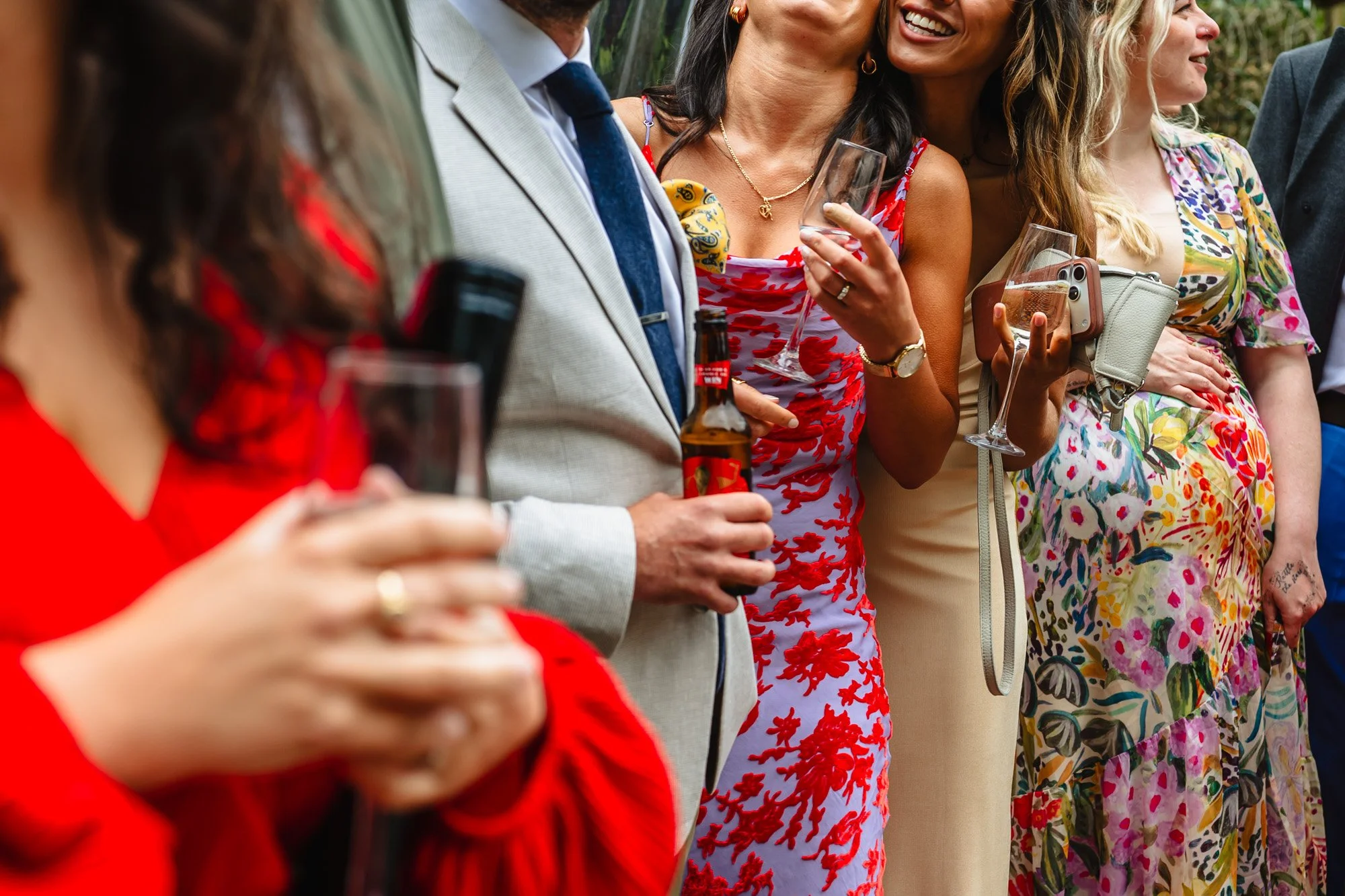 photo of 5 wedding guests whose faces are barely visible, except one who is smiling. All are holding drinks except one who is holding her pregnant tummy. Photographed by documentary wedding photographer Caroline Goosey