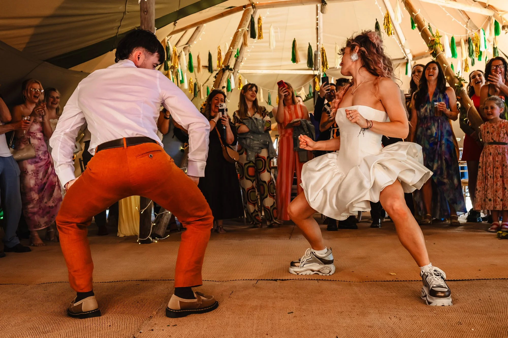 Photograph of a bride and groom dancing at their wedding in a tipi while their guests watch on, laughing and looking happy. The bride is in a super mini dress with trainers, and the groom is wearing a white shirt and orange linen trousers.