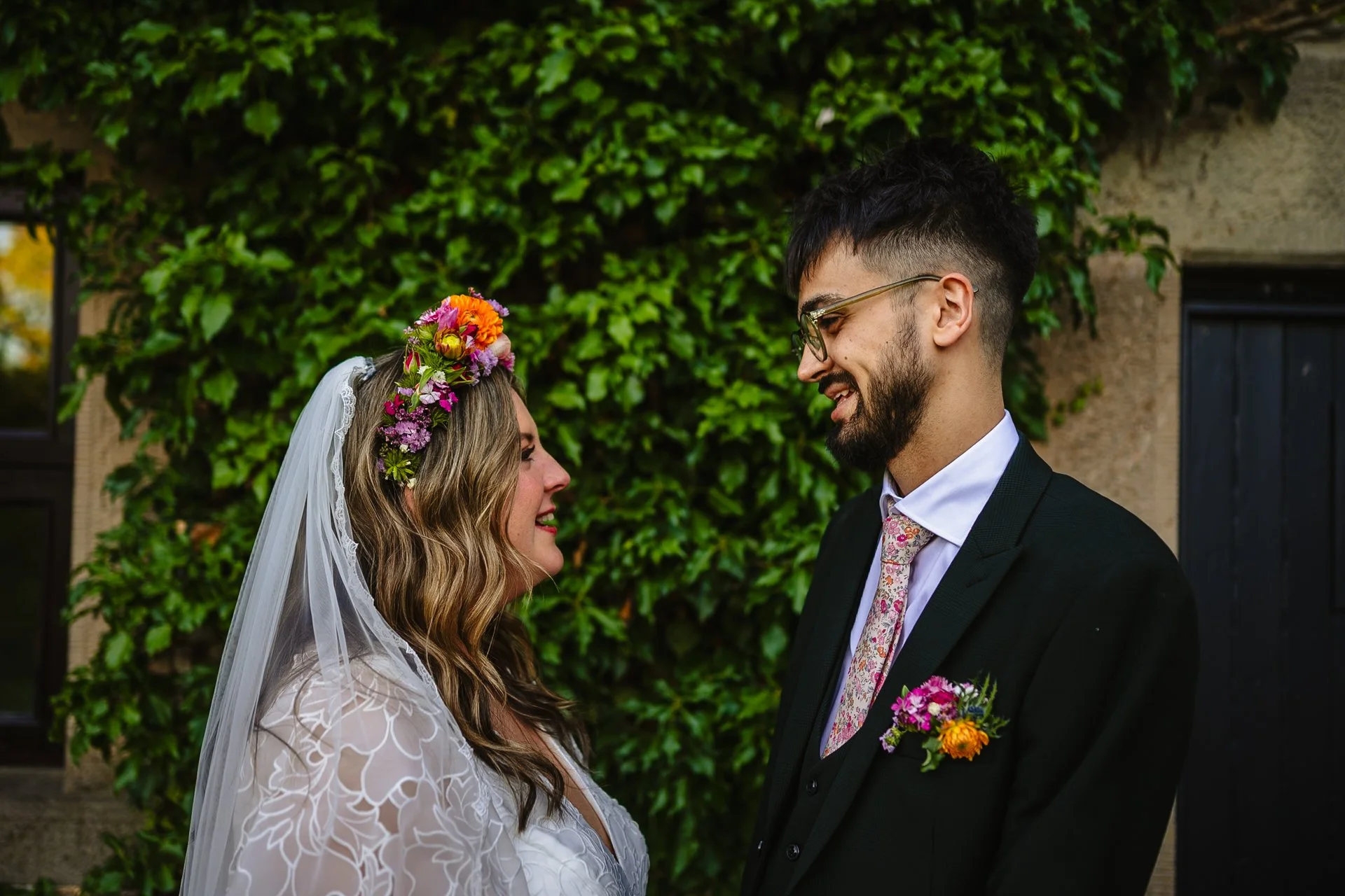A man and women face each other, smiling, in front of an ivy covered building at Peak District Holiday Barn. It is their wedding day, and she is wearing a white dress, veil and colourful flower crown. He is wearing a dark green suit with a floral tie