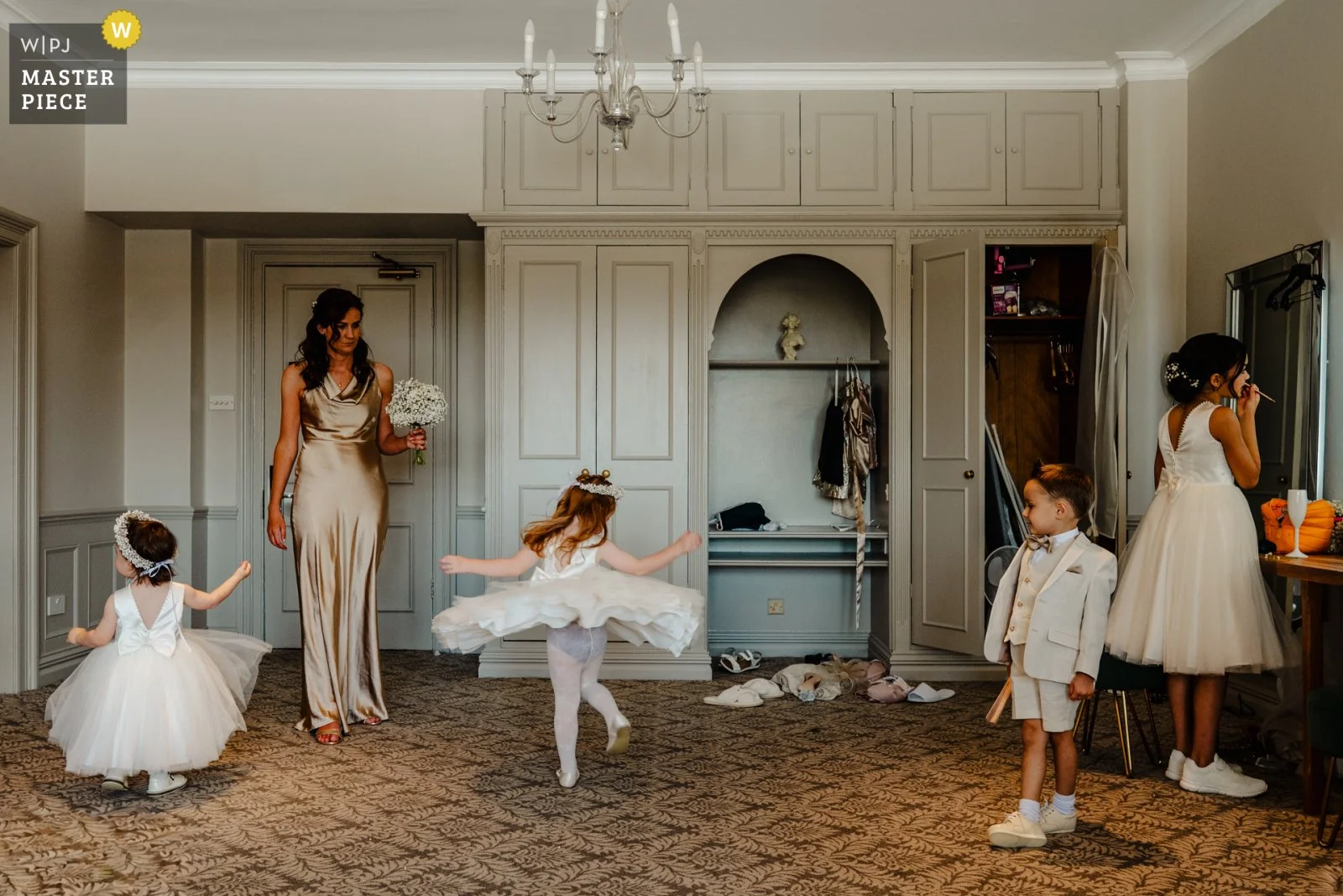 Photograph of 4 children of varying ages in smart light coloured clothing ready for a wedding ceremony. A bridesmaid in a champagne coloured satin dress is watching them and holding a bouquet of gypsophilia. The children are all amusing themselves in