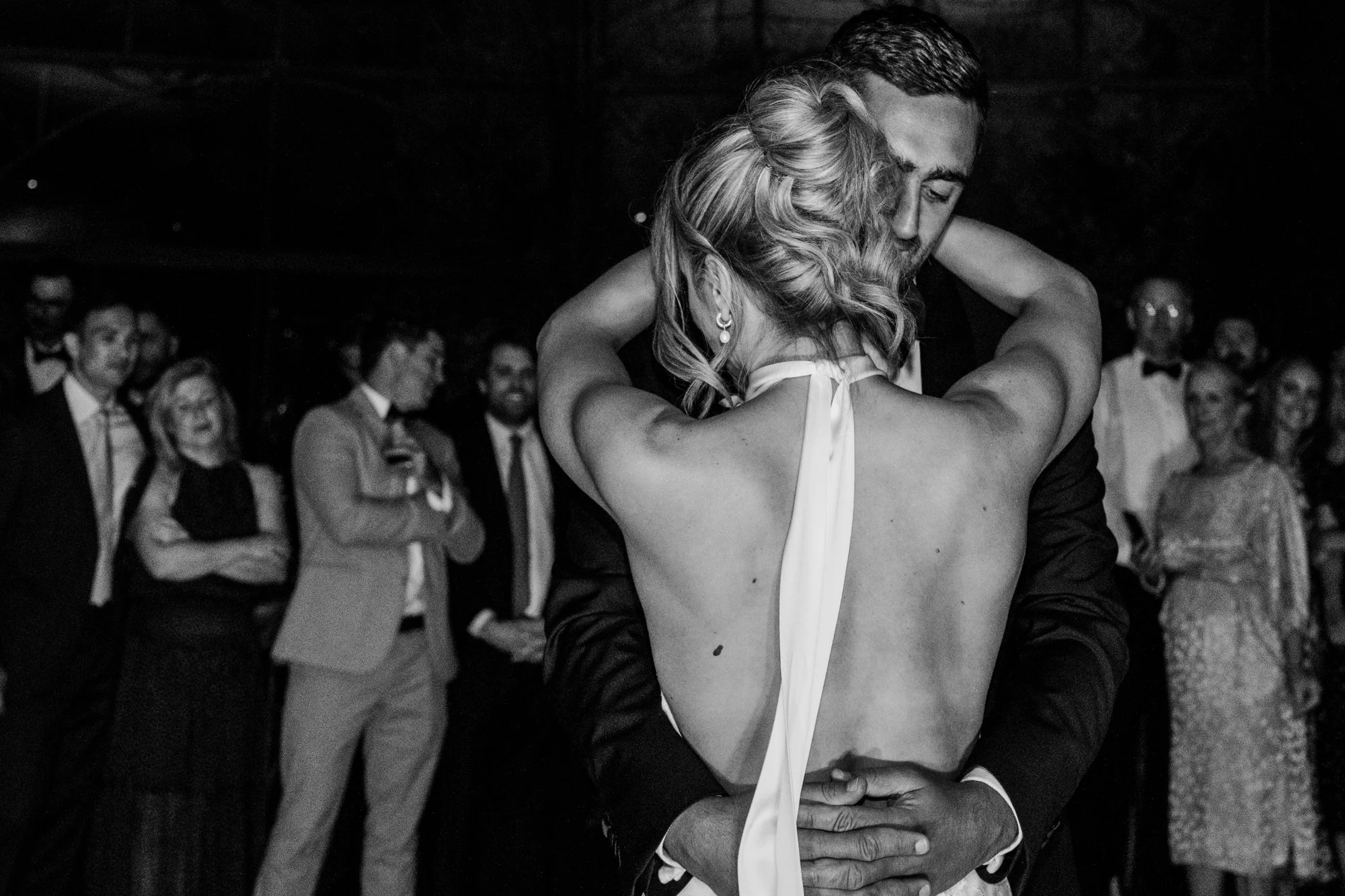 Documentary style black and white photograph of a couple dancing during their wedding. Guests are visible in the background. The couple at hugging each other. Bride wears a backless gown.