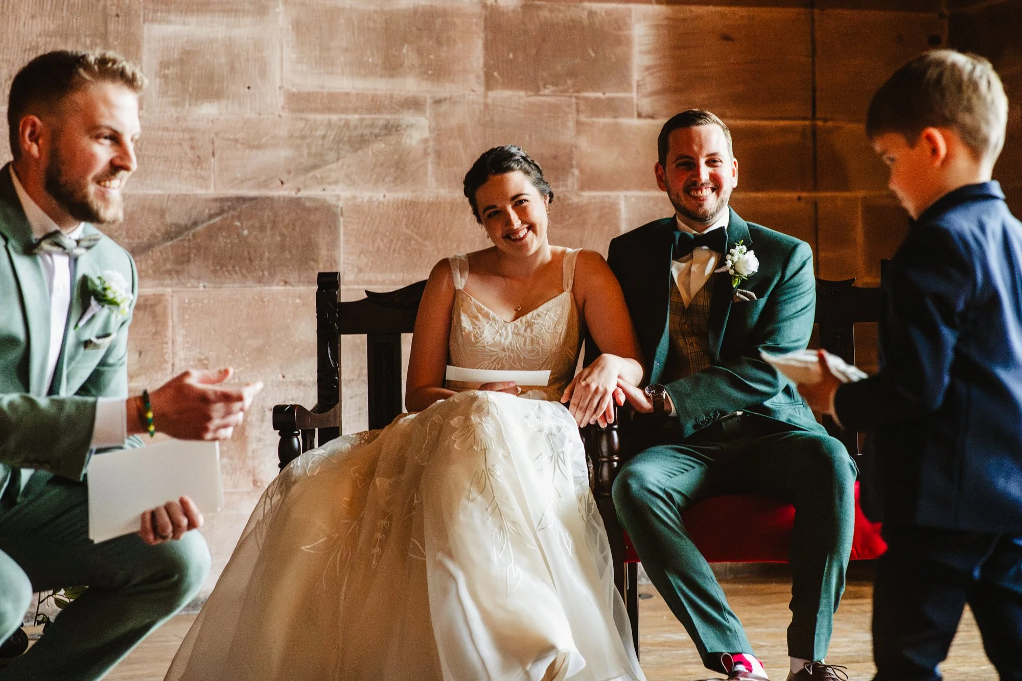 Photograph of a couple during their wedding ceremony at Peckforton Castle. The couple are sitting on chairs while a little boy delivers their rings on a cushion to his dad, who is carrying out the ceremony.