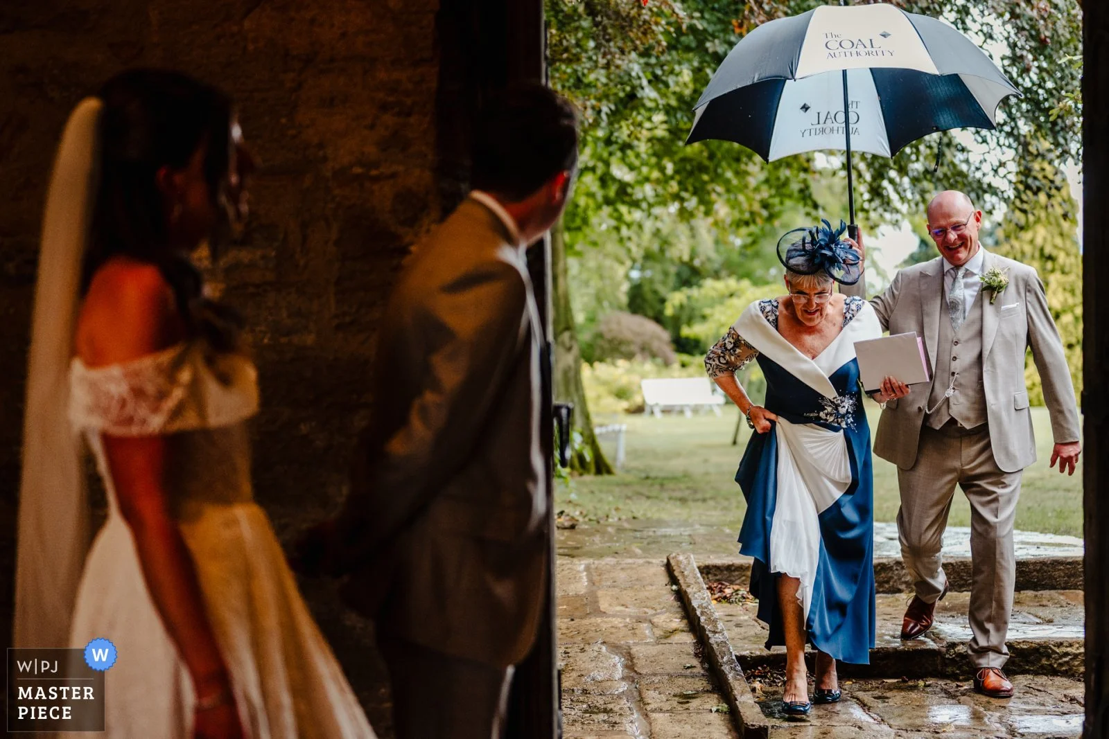 Photograph of a bride and groom waiting by an open door to greet two older wedding guests, who are walking with an umbrella. There's a WIPJ masterpiece award for documentary wedding photography in one corner.