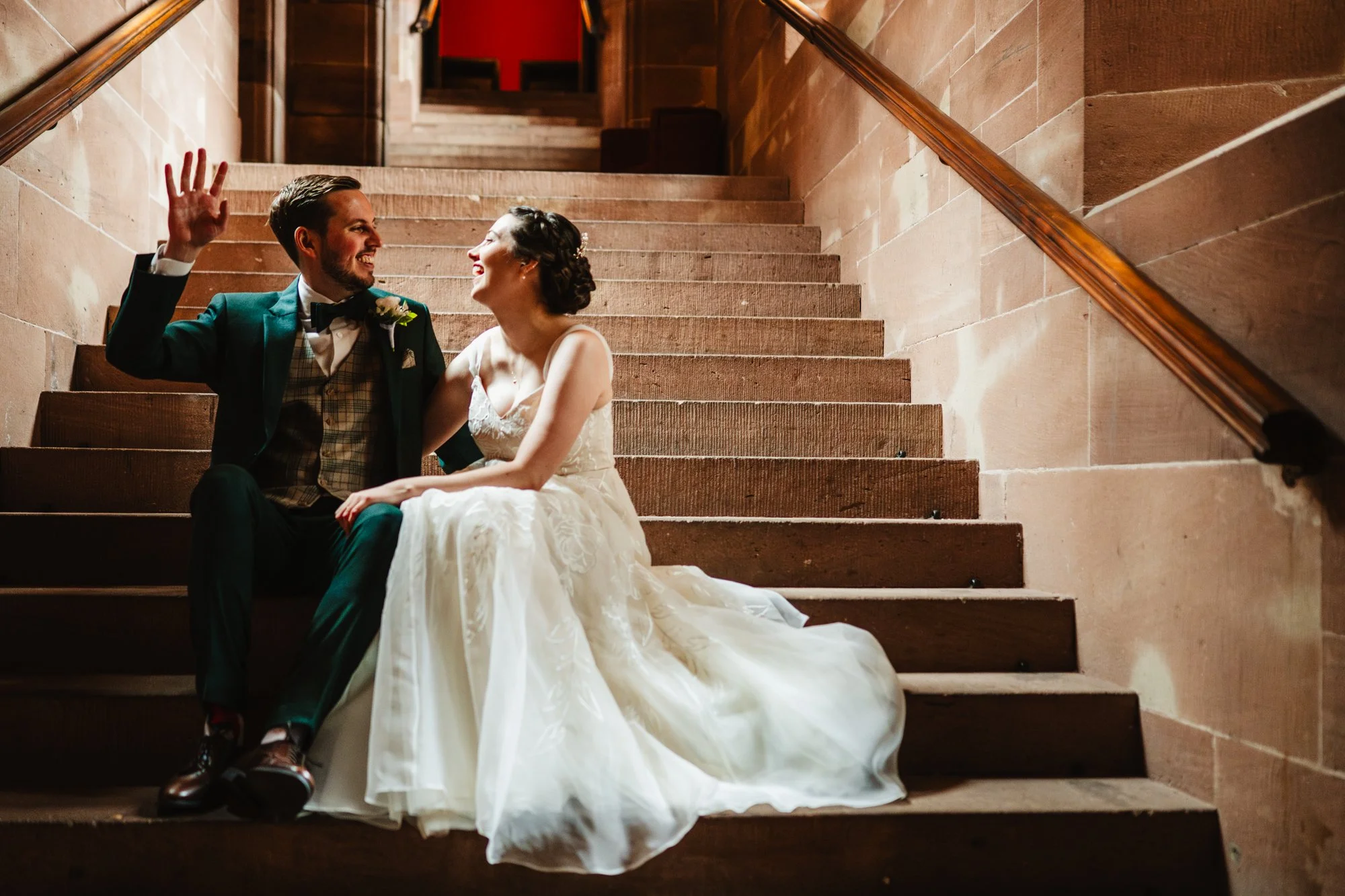 A newly married couple sit on the stone steps during their wedding at Peckforton Castle. They are smiling and talking animatedly.