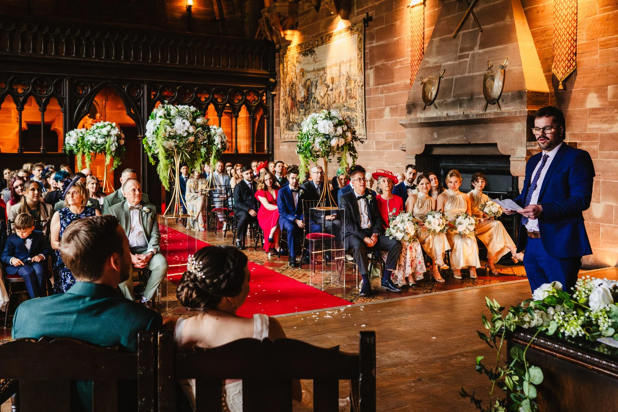 Photograph of a wedding ceremony in the Great Hall at Peckforton Castle in Cheshire. A man in a blue suit is reading while looking at the couple, who are seated on chairs in the foreground. Their guests watch from their seated position.
