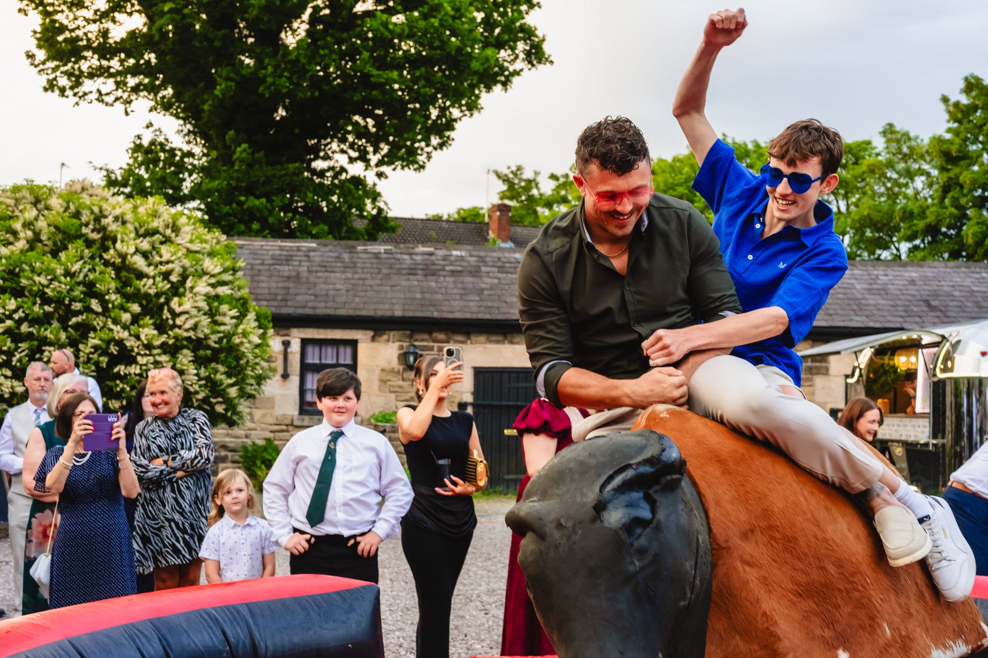 Two men ride a rodeo bull together in the car park at Lost Wirral wedding venue. They are both wearing colourful heart shaped sunglasses and grinning. There's a crowd of people watching.