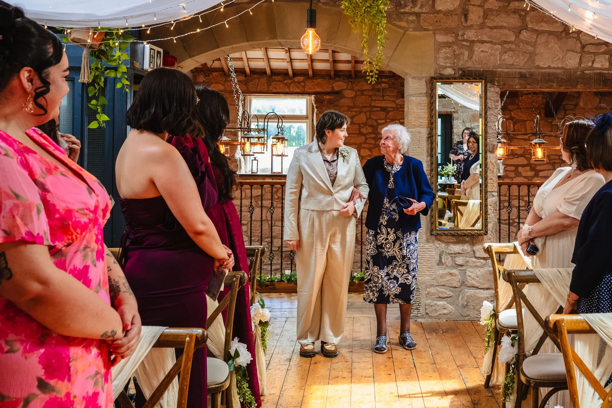 photograph of a bride in a silk suit and short hair walking down the aisle with her grandma at Lost wedding venue on the wirral